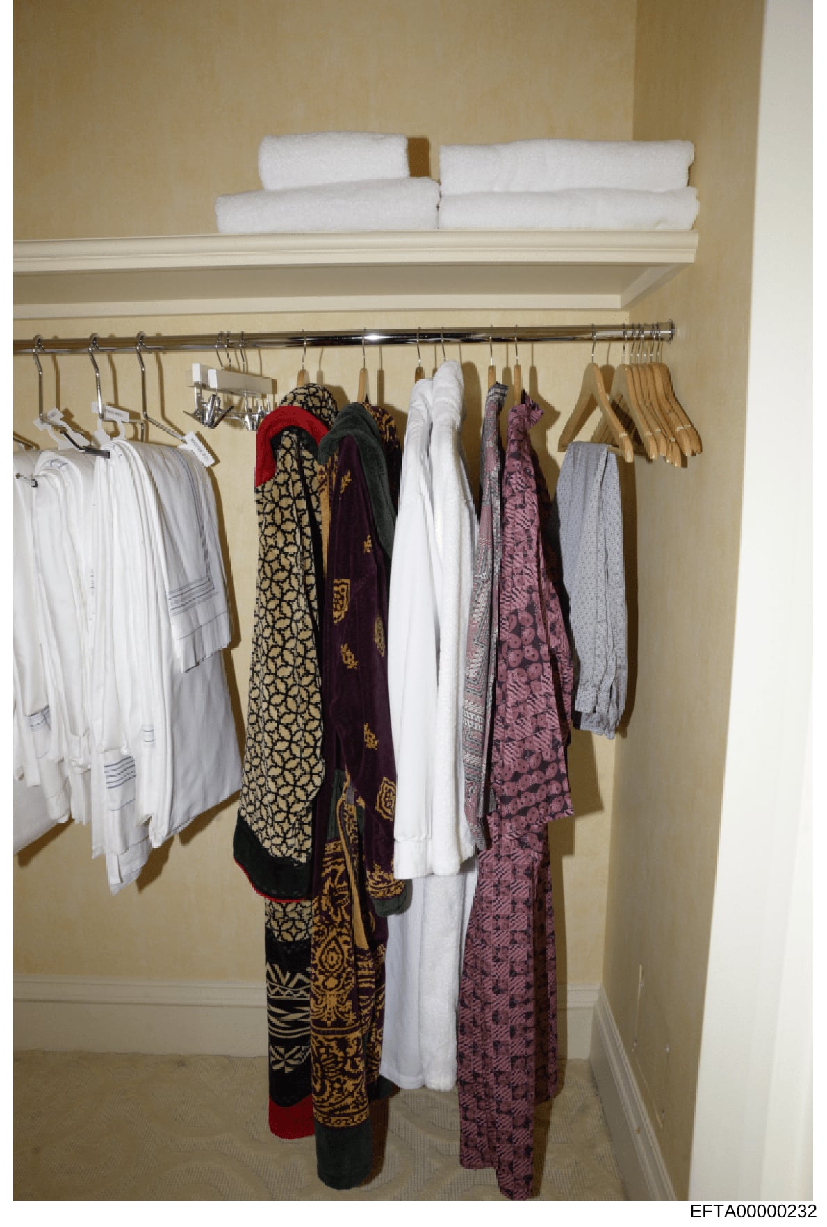 This photograph shows the interior of a bathroom or bedroom closet containing folded towels on an upper shelf, wooden hangers with various clothing items including white garments, patterned scarves or wraps in black and gold, burgundy, and pink tones