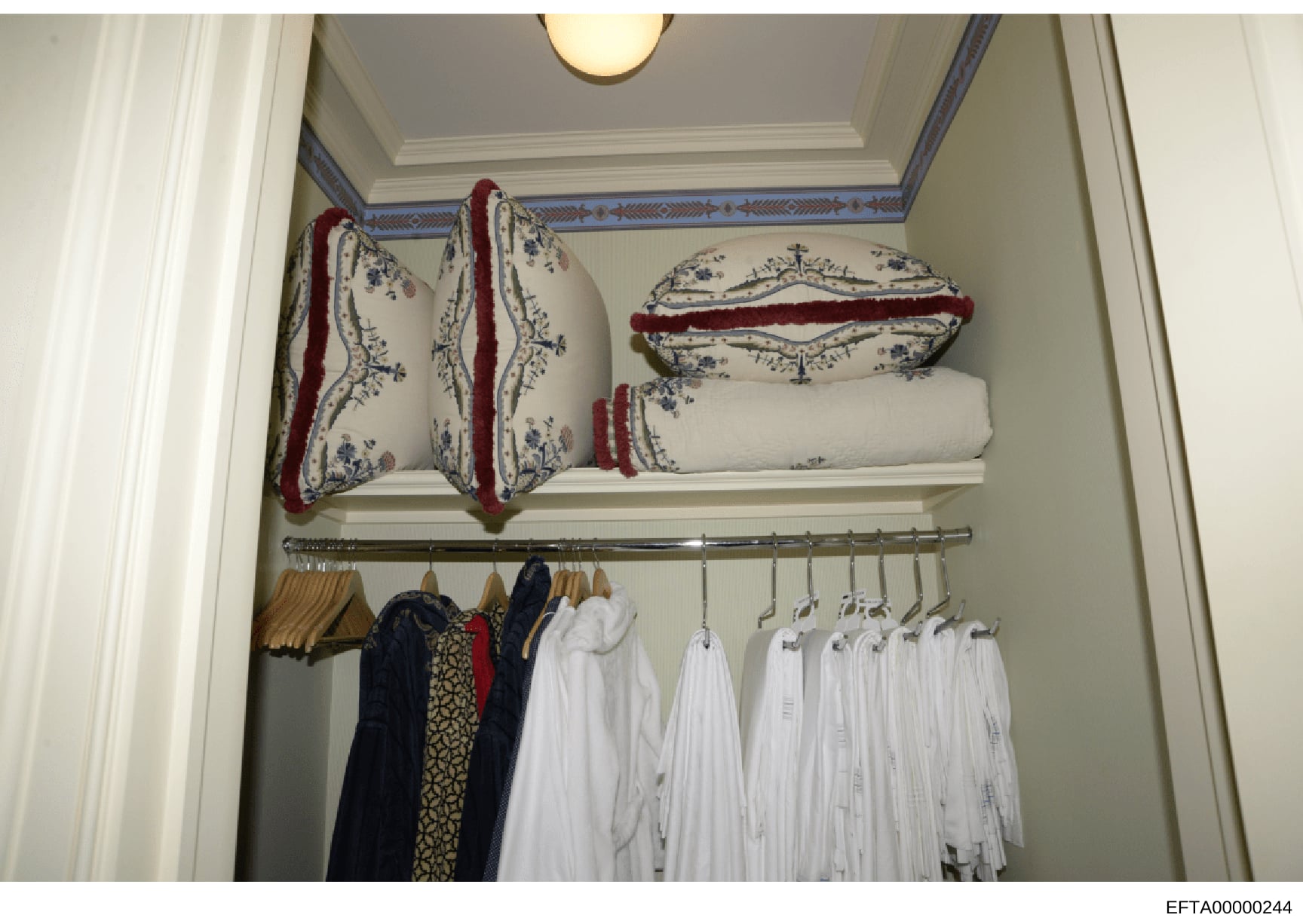 This is a photograph of an organized closet interior, likely from one of Jeffrey Epstein's residences. The image shows shelving with decorative pillows and linens with red velvet trim and blue floral patterns on the upper shelf, and a clothing rod be