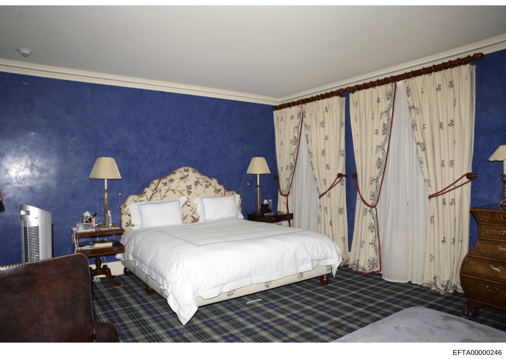 This is a photograph of a luxuriously appointed bedroom featuring deep blue walls, a decorative upholstered headboard, white bedding, cream-colored patterned curtains with burgundy tie-backs, wooden furniture, and a plaid carpet. The photograph appea