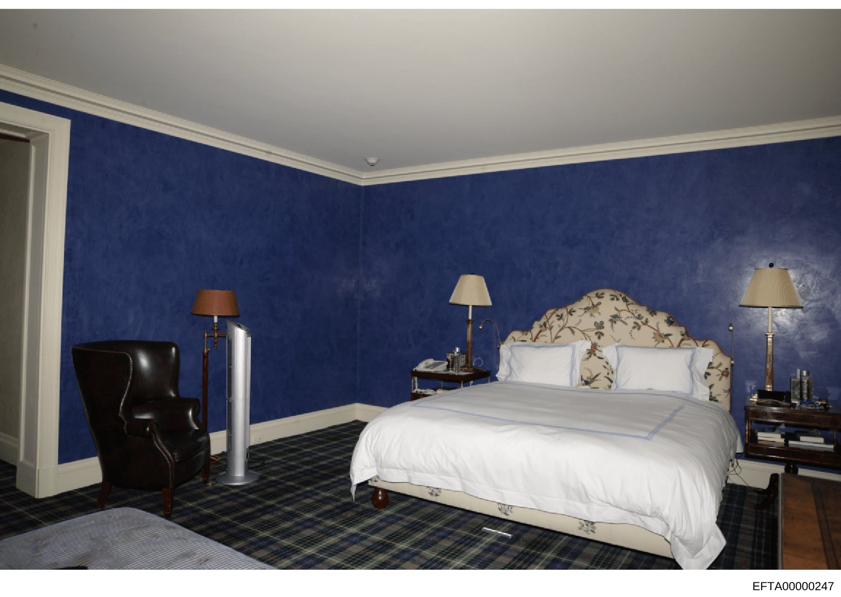 This is a photograph of an interior bedroom space featuring distinctive blue velvet-textured wall coverings, a decorative bed with floral upholstered headboard, and period furnishings. The room includes a black leather chair, multiple table lamps, an