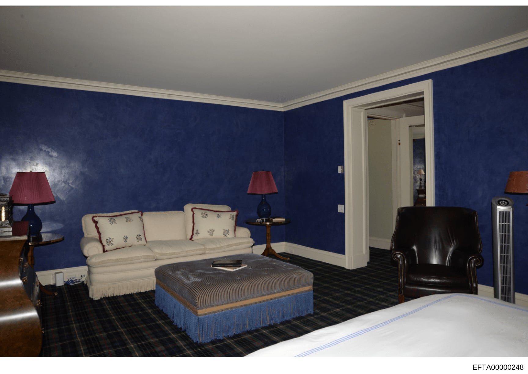 This is a photograph of an elegantly furnished bedroom featuring deep blue walls, cream-colored seating furniture, and formal décor. The room contains a cream sofa with decorative pillows, a black leather armchair, a striped ottoman with blue skirt, 