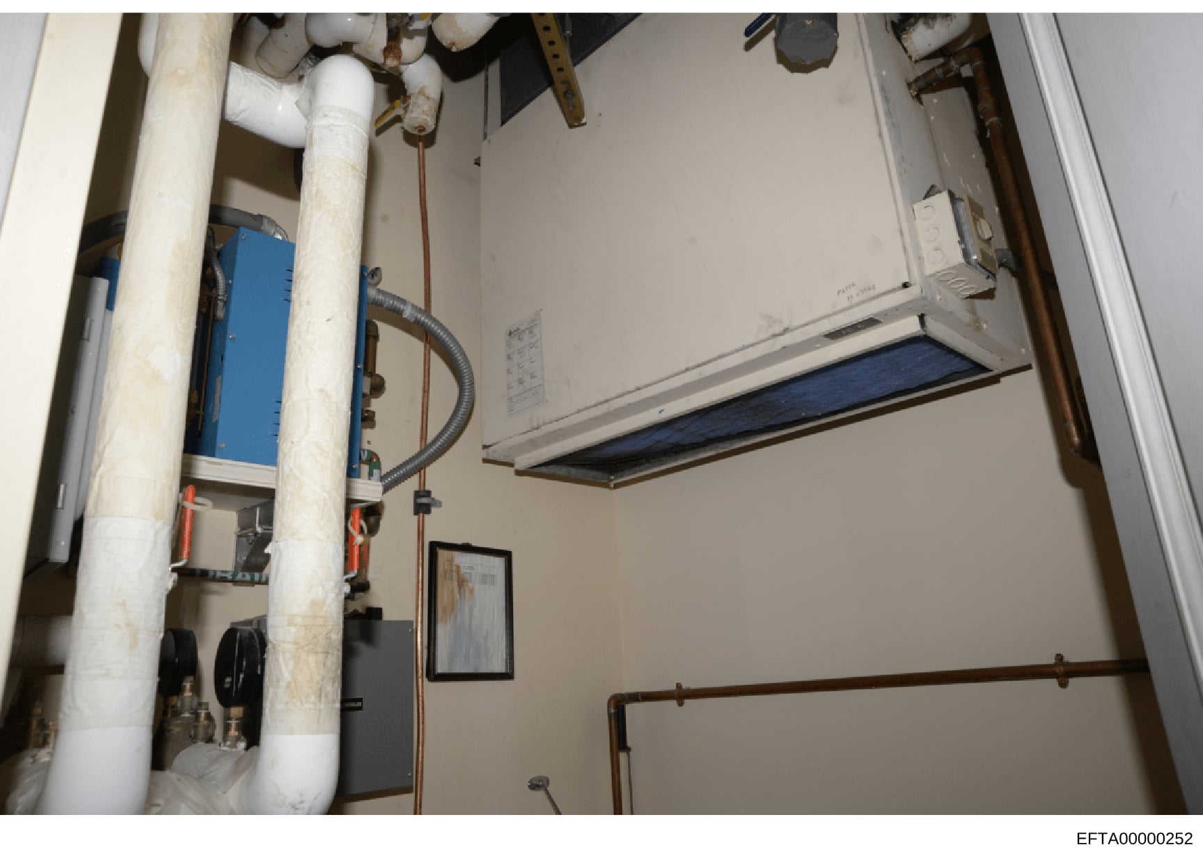 This photograph shows the interior of a utility or mechanical room, likely from one of Epstein's properties. The image captures various building systems including PVC piping, a blue water heater unit, copper piping, and ceiling-mounted equipment. The