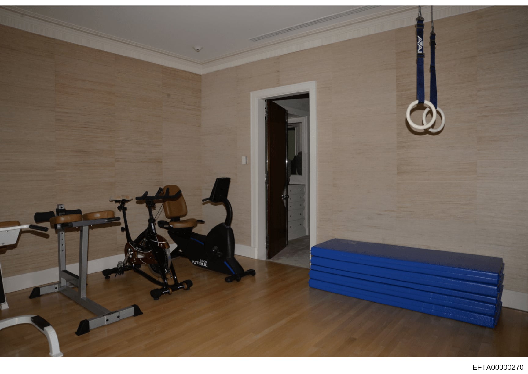 This is an evidence photograph showing a private residential gymnasium containing various exercise equipment including stationary bikes, weight benches, gymnastics mats, and suspended rings. The photo documents the interior of what appears to be a hi