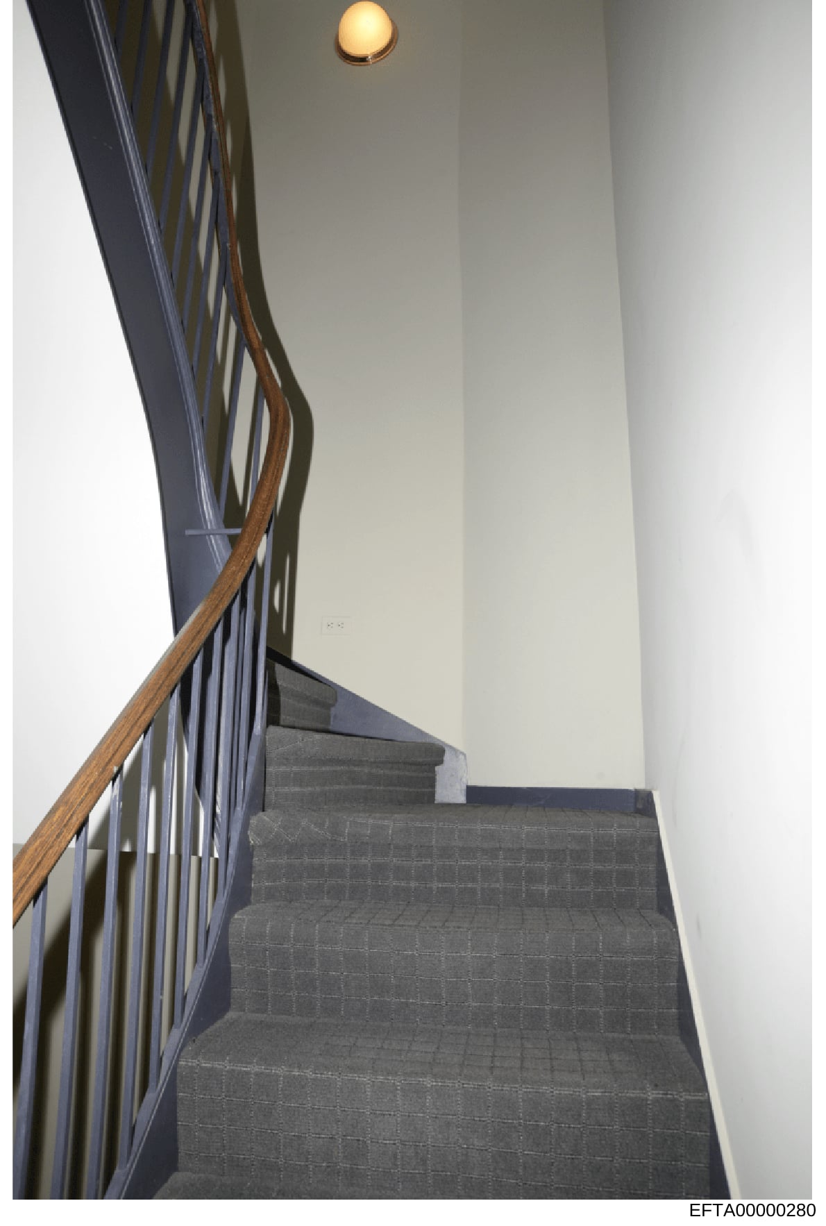 This is a photograph of an interior stairwell featuring gray carpeted stairs descending downward, a wooden handrail on the left side, white walls, and a round ceiling light fixture. The image appears to be documentation of a property interior, likely