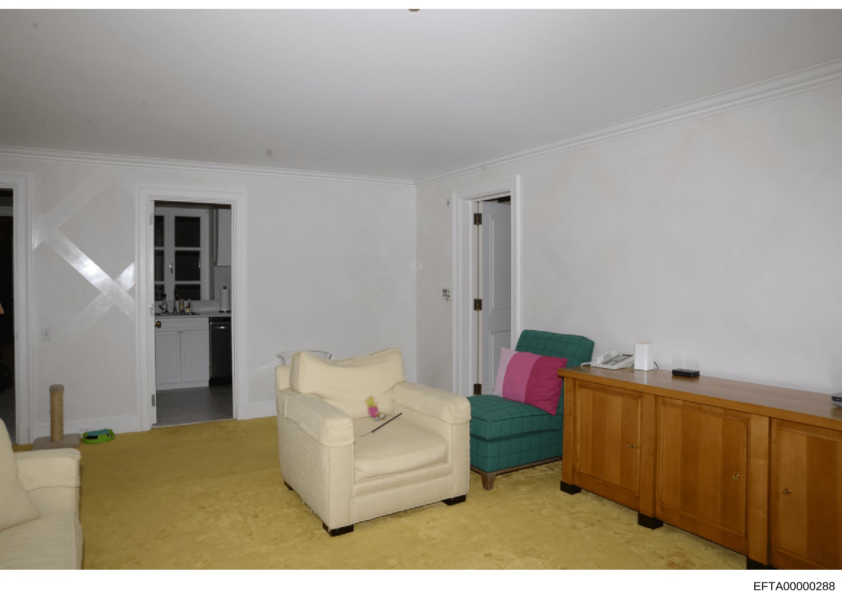 This is a photograph of a residential living room, showing furniture placement and interior design elements. The room features cream-colored walls, a yellow carpet, upholstered seating including a cream armchair and green/pink sectional pieces, woode