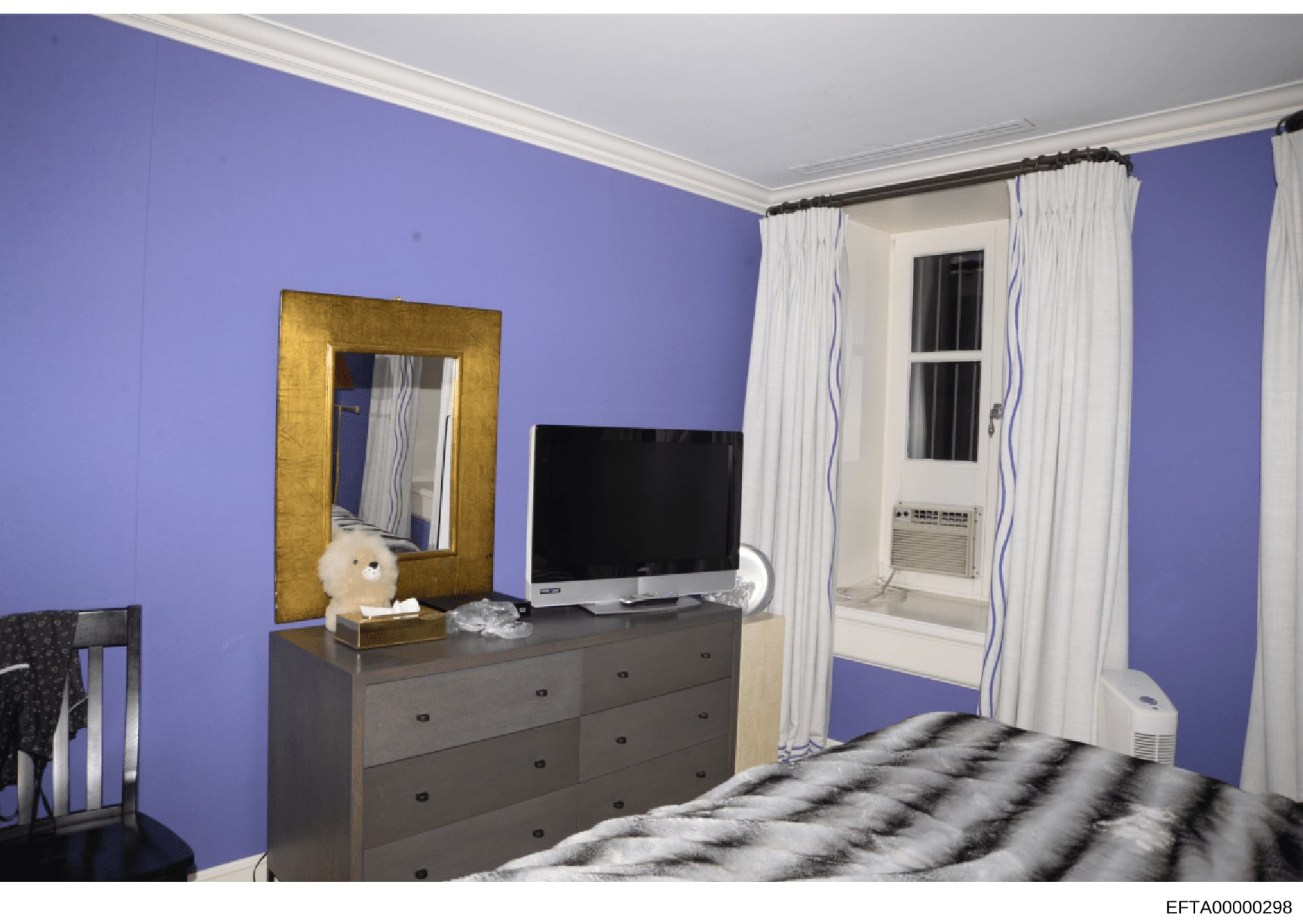 This is a photograph of a bedroom interior featuring distinctive purple-painted walls, a gray dresser with television, a gold-framed mirror, and a bed with patterned bedding. The room includes a window with white curtains, an air conditioning unit, a