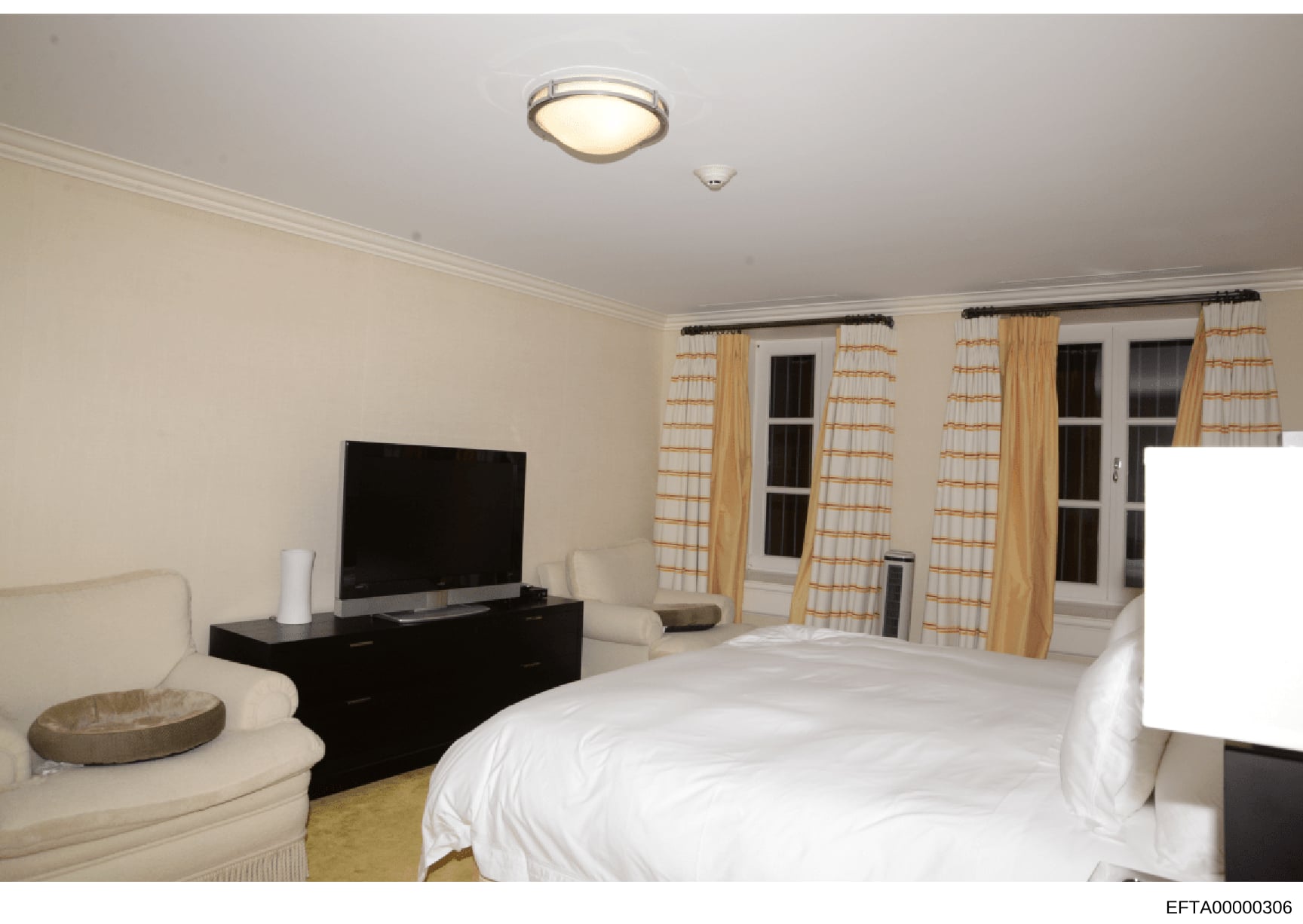 This is a photograph of a luxury bedroom interior featuring a king-sized bed, entertainment system, and floor-to-ceiling windows with custom drapes. The room displays high-end furnishings and décor consistent with a wealthy property. The image appear