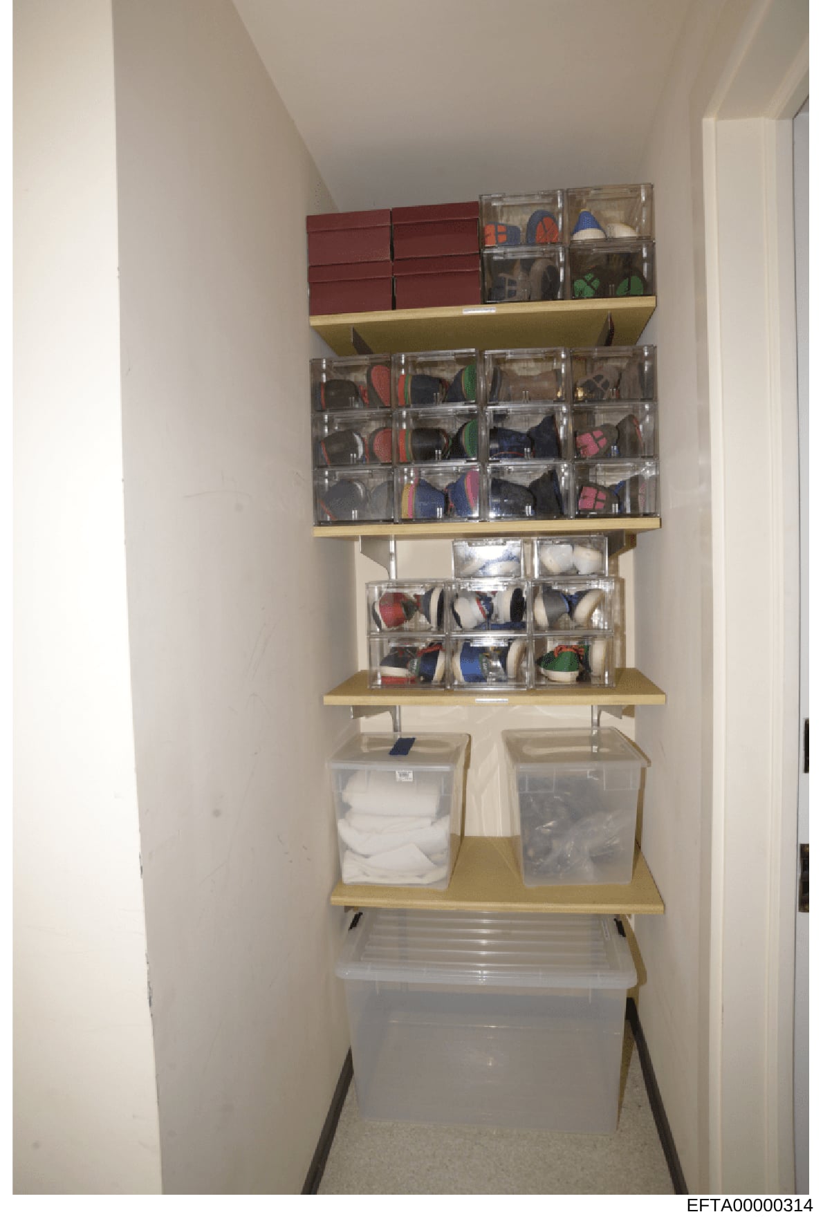 This photograph shows a well-organized storage closet featuring wall-mounted shelving units with multiple clear plastic containers holding various items. The closet contains burgundy storage boxes at the top, several shelves with labeled clear contai
