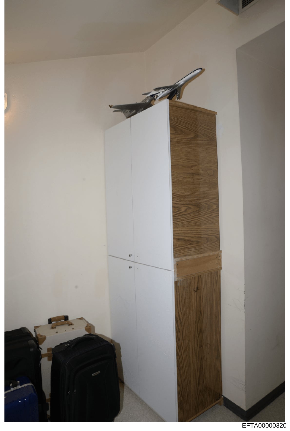 This is an evidence photograph showing the interior of a closet or small room containing a tall white and wood-grain cabinet/wardrobe, luggage items on the floor, and a model aircraft positioned on top of the cabinet. The photo is part of the Epstein
