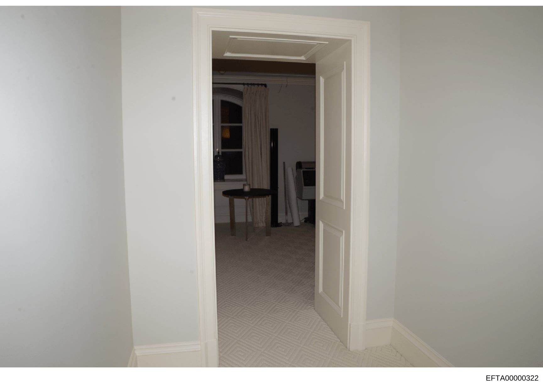 This photograph depicts an interior hallway or corridor with white walls and a geometric patterned carpet. The doorway opens into an adjacent room with darker walls, where furniture including chairs and what appears to be stored materials are visible