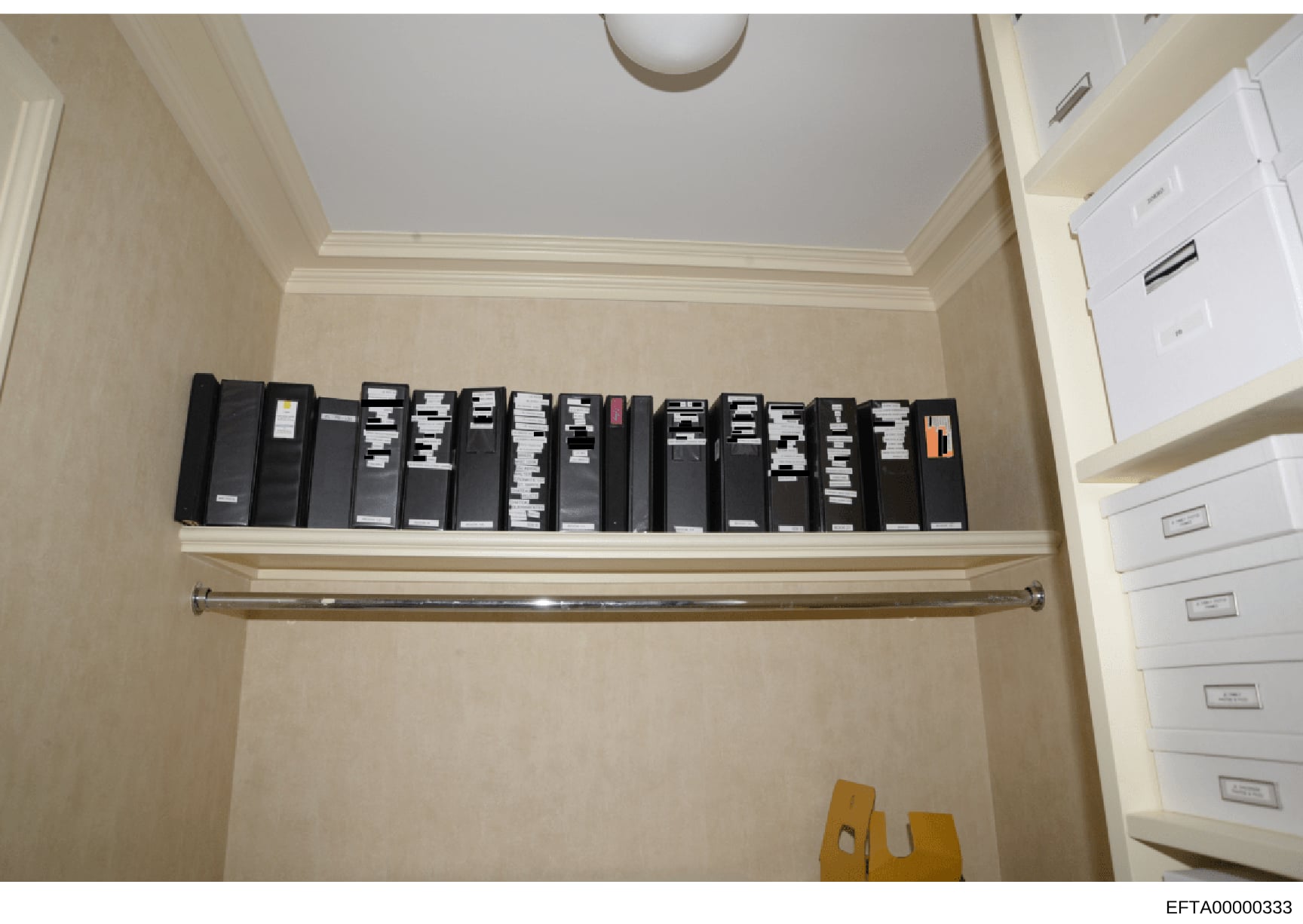 This is an evidence photograph showing a closet or storage area containing approximately 16-18 black binders arranged on a shelf, along with white filing cabinets visible on the right side. The binders appear to be labeled or indexed, suggesting orga