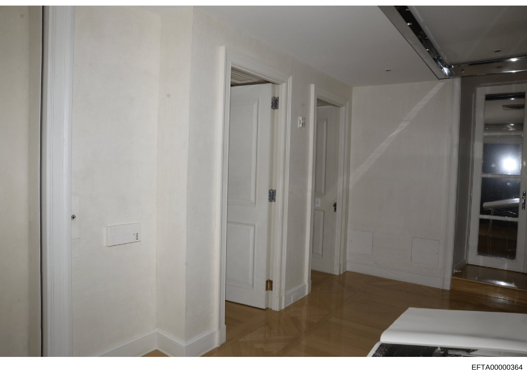 This is a photograph of an interior hallway within a residential or commercial property, showing white painted walls, multiple white doors with brass hardware, wooden flooring, recessed lighting, and what appears to be a mirror or reflective surface