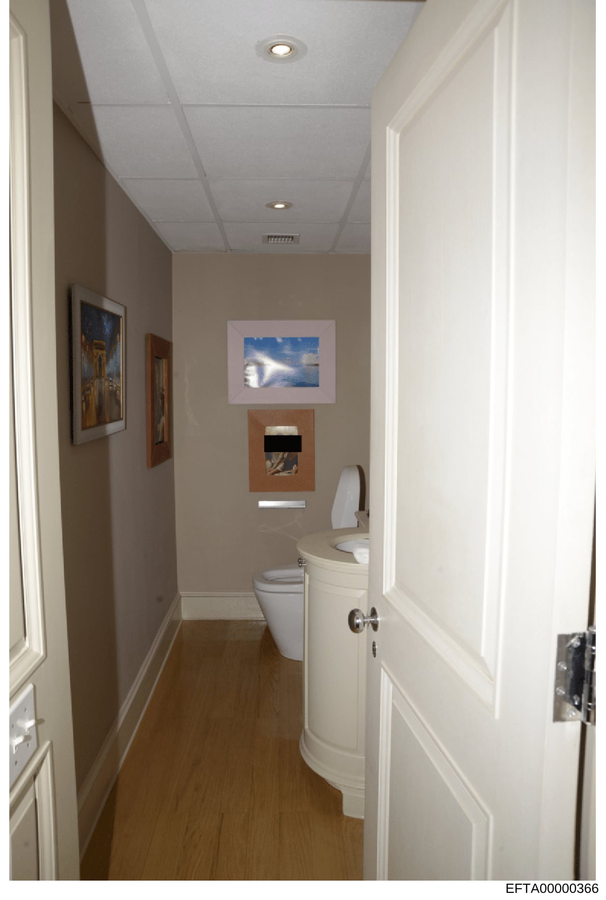 This is a photograph of a narrow, well-maintained bathroom featuring beige and white décor, wood flooring, and multiple framed artworks on the walls. The image appears to be documentation of a property interior, likely taken during an investigation o