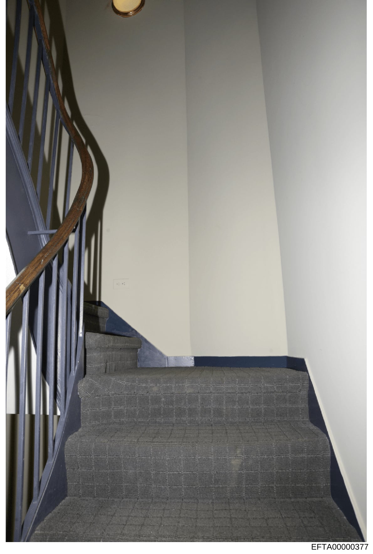 This is a photograph of an interior staircase, likely from one of Epstein's properties documented during the investigation. The image shows a flight of stairs with gray carpet, a wooden handrail with metal balusters, and white walls. The photo appear