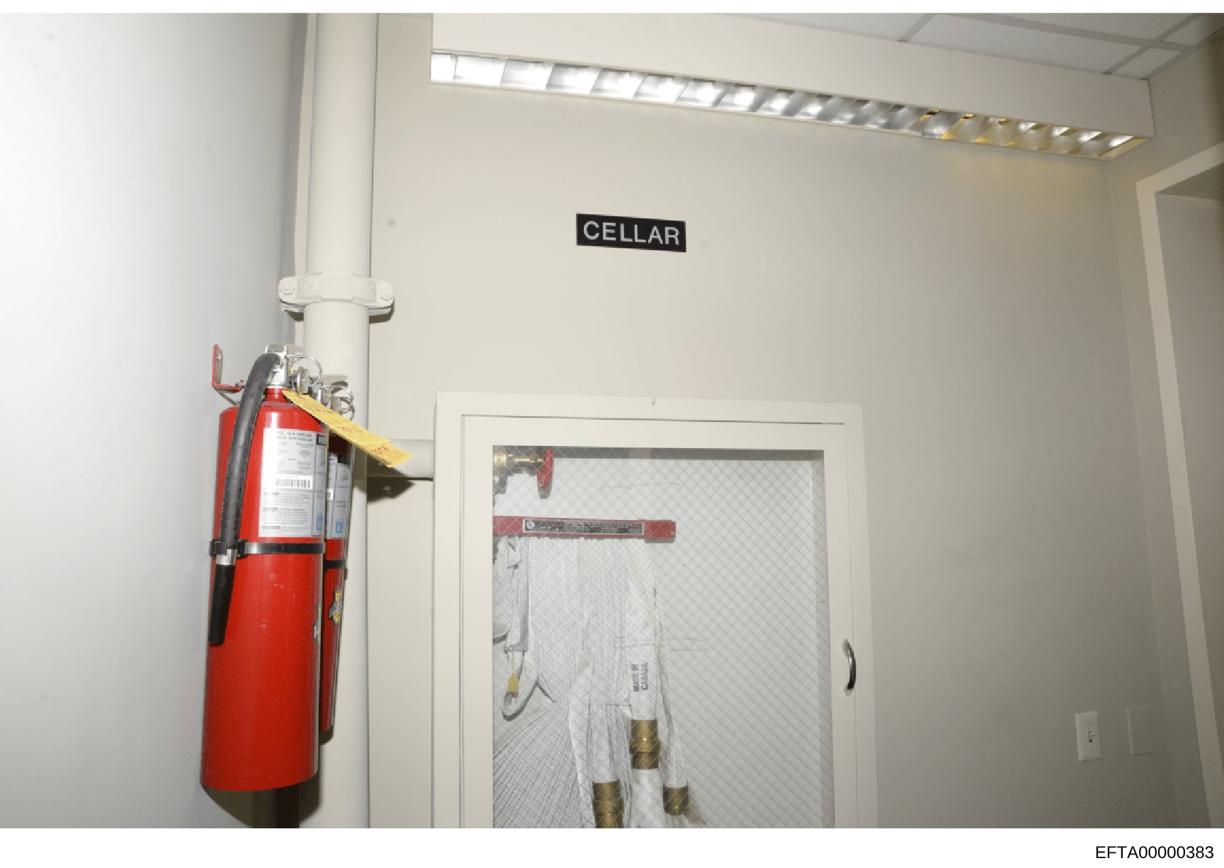 This is a photograph of a cellar room showing basic infrastructure and safety equipment. The image depicts a white-walled basement space with a fire extinguisher mounted on the left wall, fluorescent lighting, and what appears to be storage or utilit