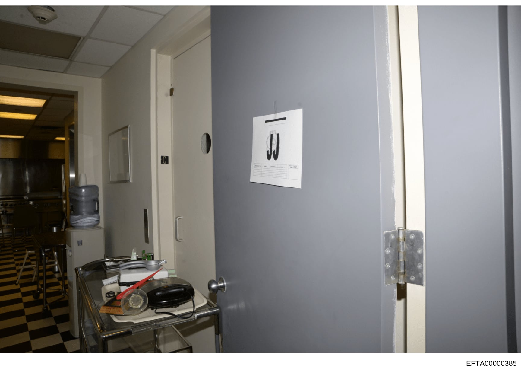 This evidence photograph shows an interior room with light blue/gray walls, fluorescent ceiling, and a metal utility table. A white paper with initials 'JJ' is prominently displayed on the wall, suggesting this may be an office, residence, or facilit