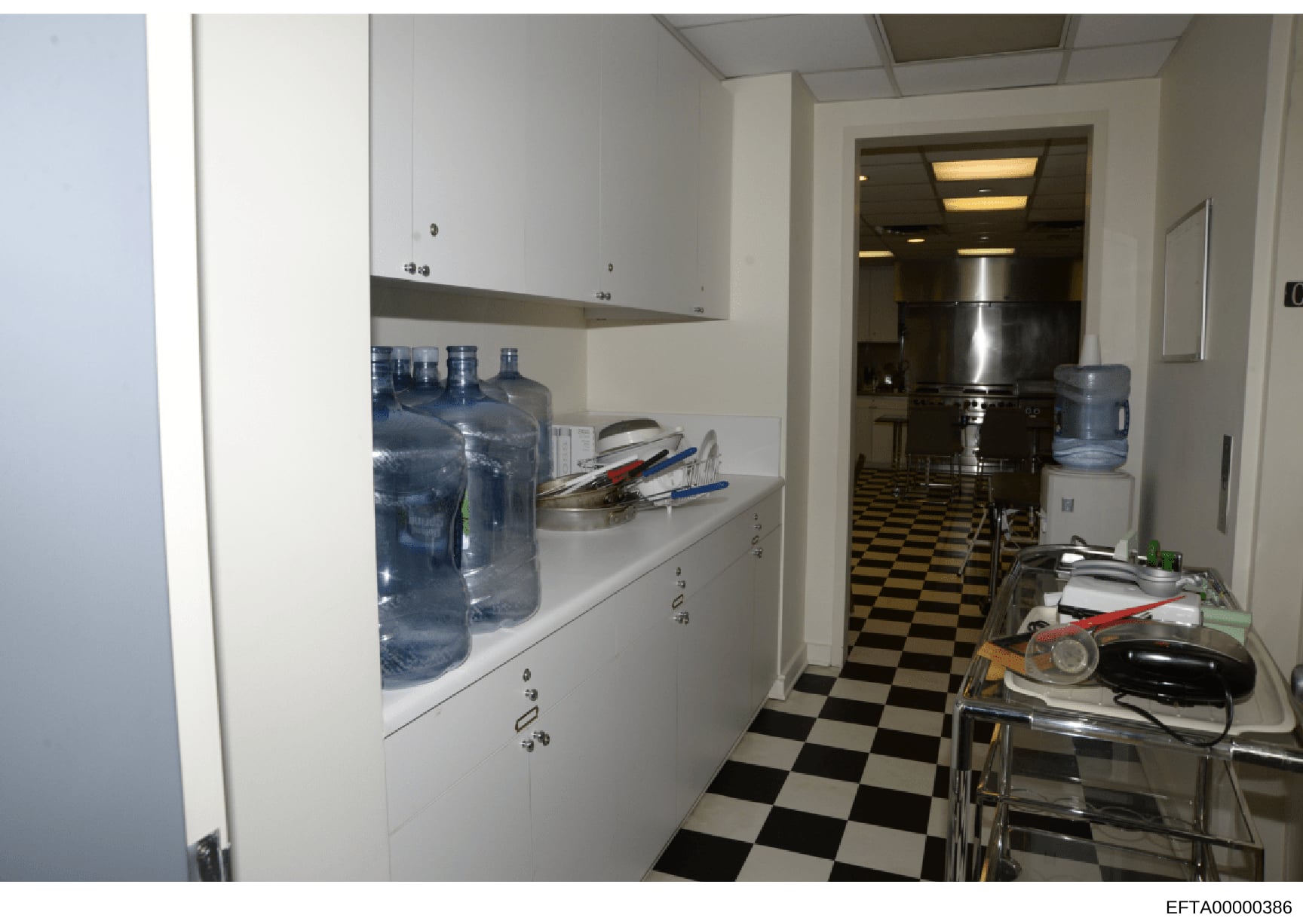 This is a photograph of what appears to be a staff or service kitchen area, featuring white cabinetry, multiple large water bottles, kitchen utensils, and a black-and-white checkerboard floor pattern. The image was taken as part of the evidence docum