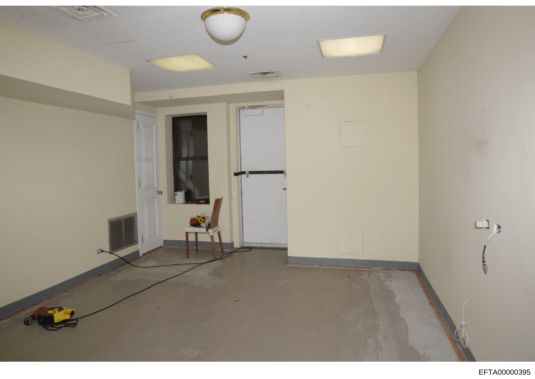 This is a photograph of an empty commercial interior space in the process of renovation or construction. The room features cream-colored walls, fluorescent ceiling fixtures, and concrete flooring with protective tape marking borders. A single wooden