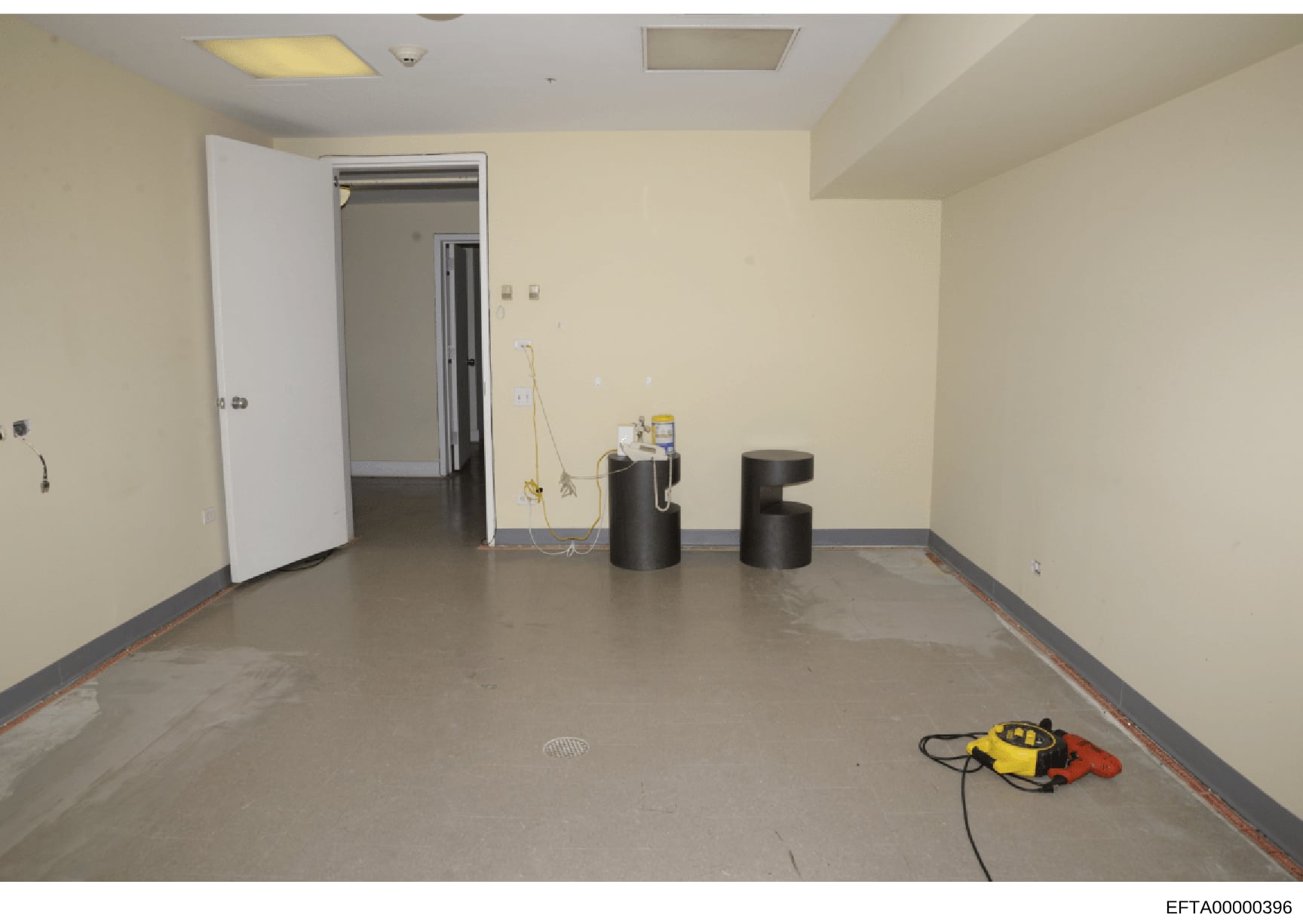 This photograph documents an empty interior room with cream-colored walls, recessed ceiling lights, and concrete flooring. The space appears to be under renovation or preparation, with electrical cords, construction equipment (yellow and red power to