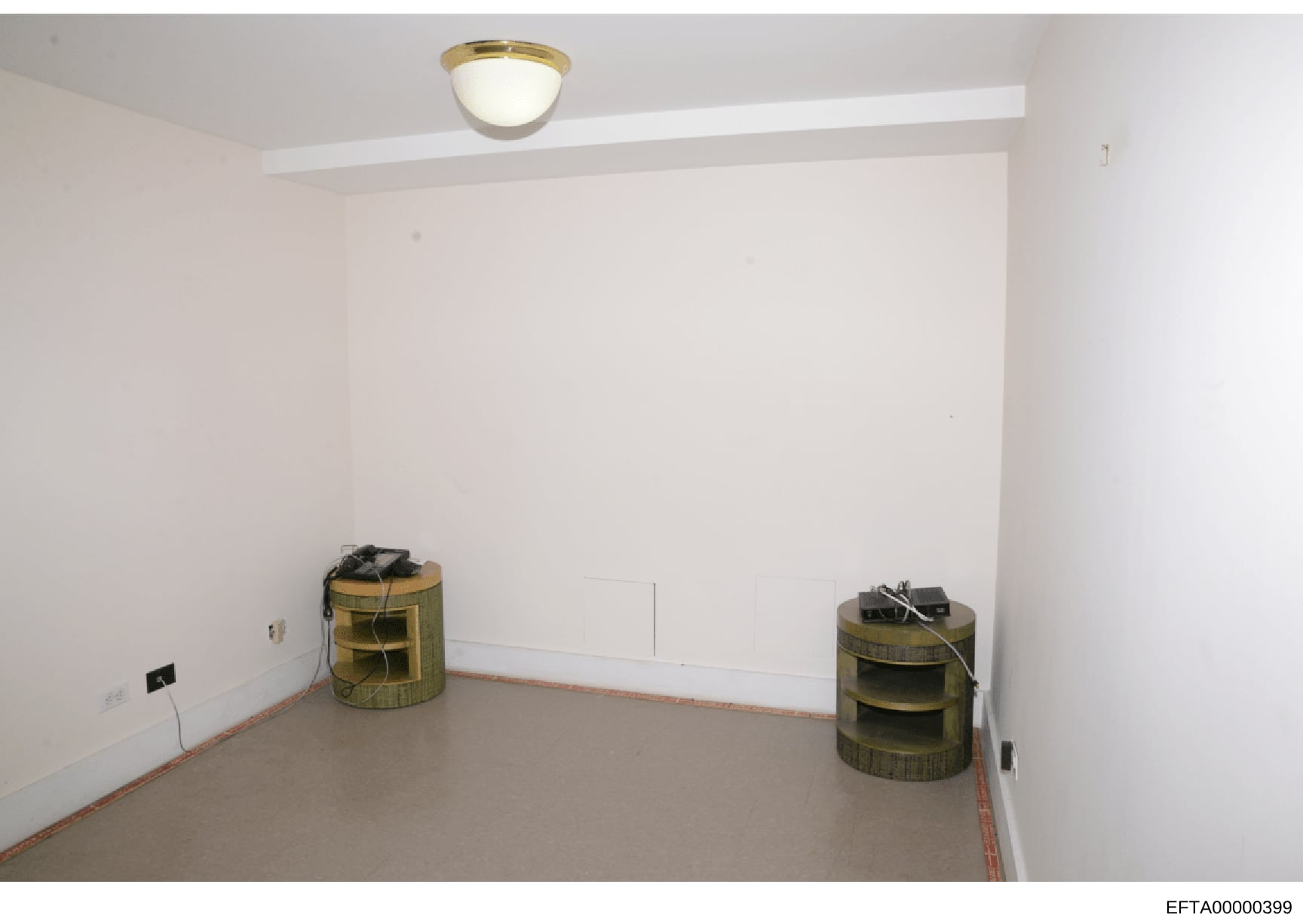 This is a photograph of an empty interior room, likely from an Epstein property during a federal investigation. The room shows white walls, a concrete floor with red base trim, a ceiling-mounted light fixture, and two cylindrical woven storage contai