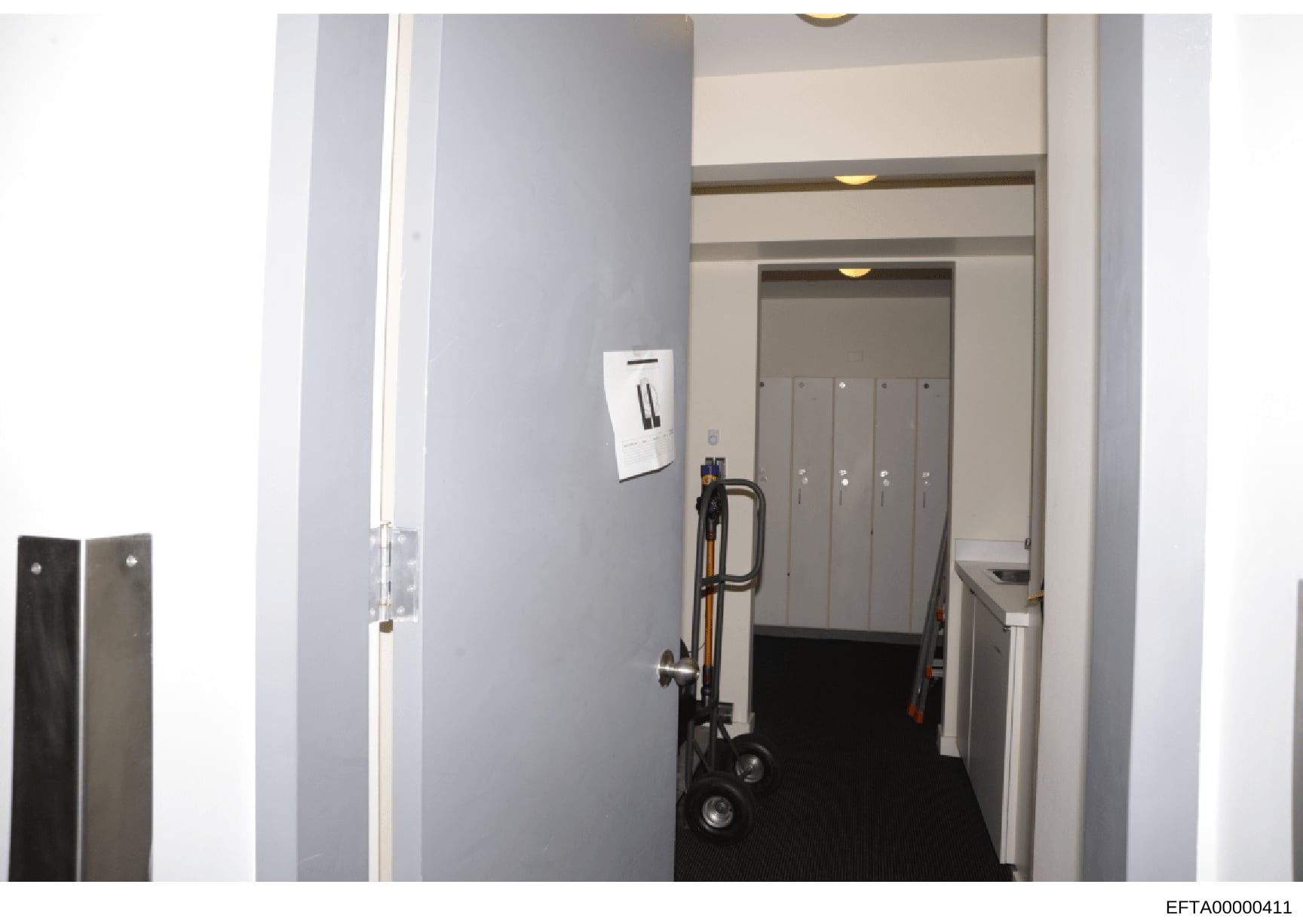This photograph depicts an interior hallway or corridor space, showing built-in storage lockers, a light switch panel, recessed ceiling lighting, and a hand truck or dolly positioned in the passageway. The image appears to be part of property documen