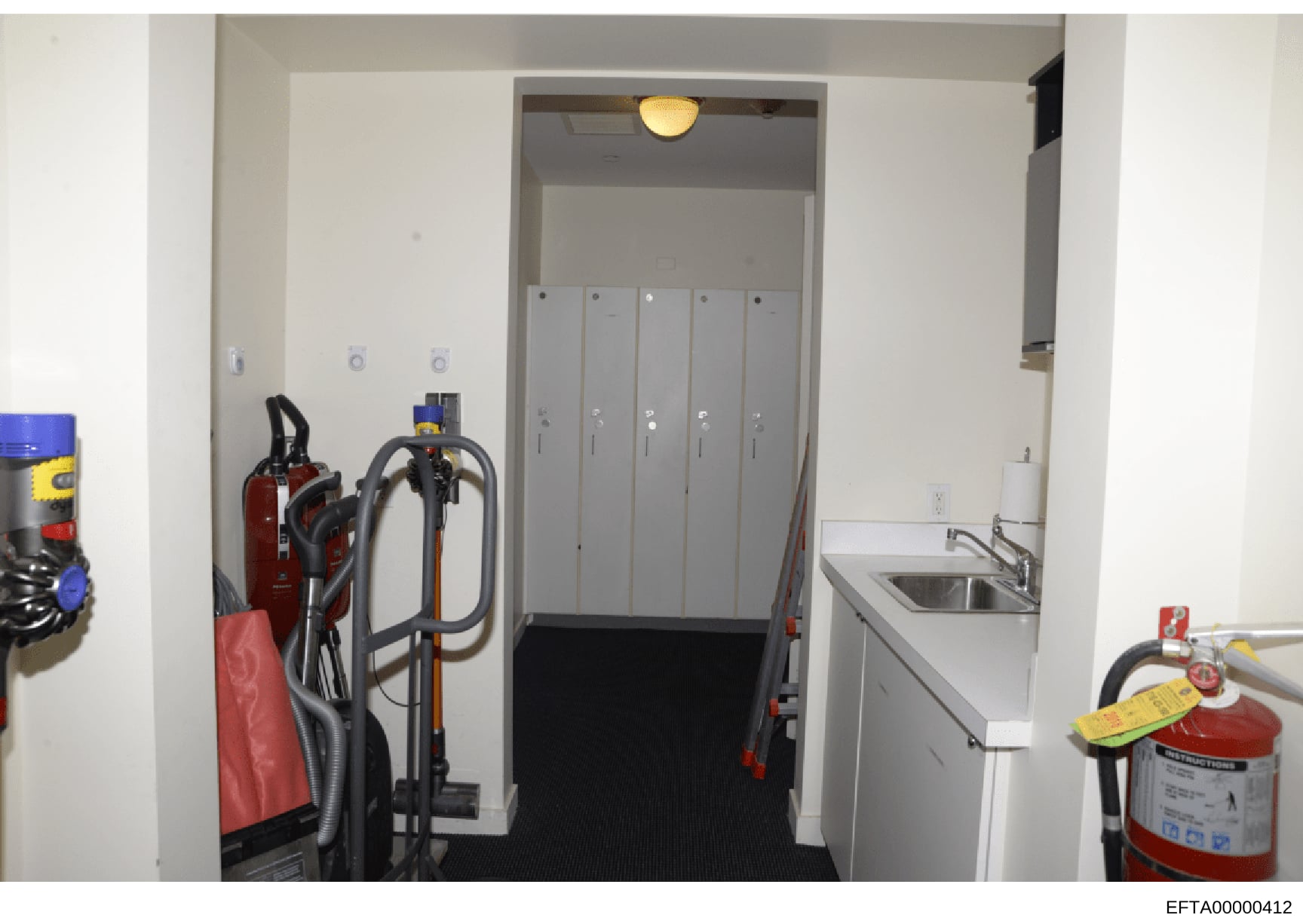 This is a photograph of an interior hallway or utility corridor showing maintenance and safety equipment including fire extinguishers, cleaning equipment, and an elevator. The space appears to be in a commercial or residential building. The image is 