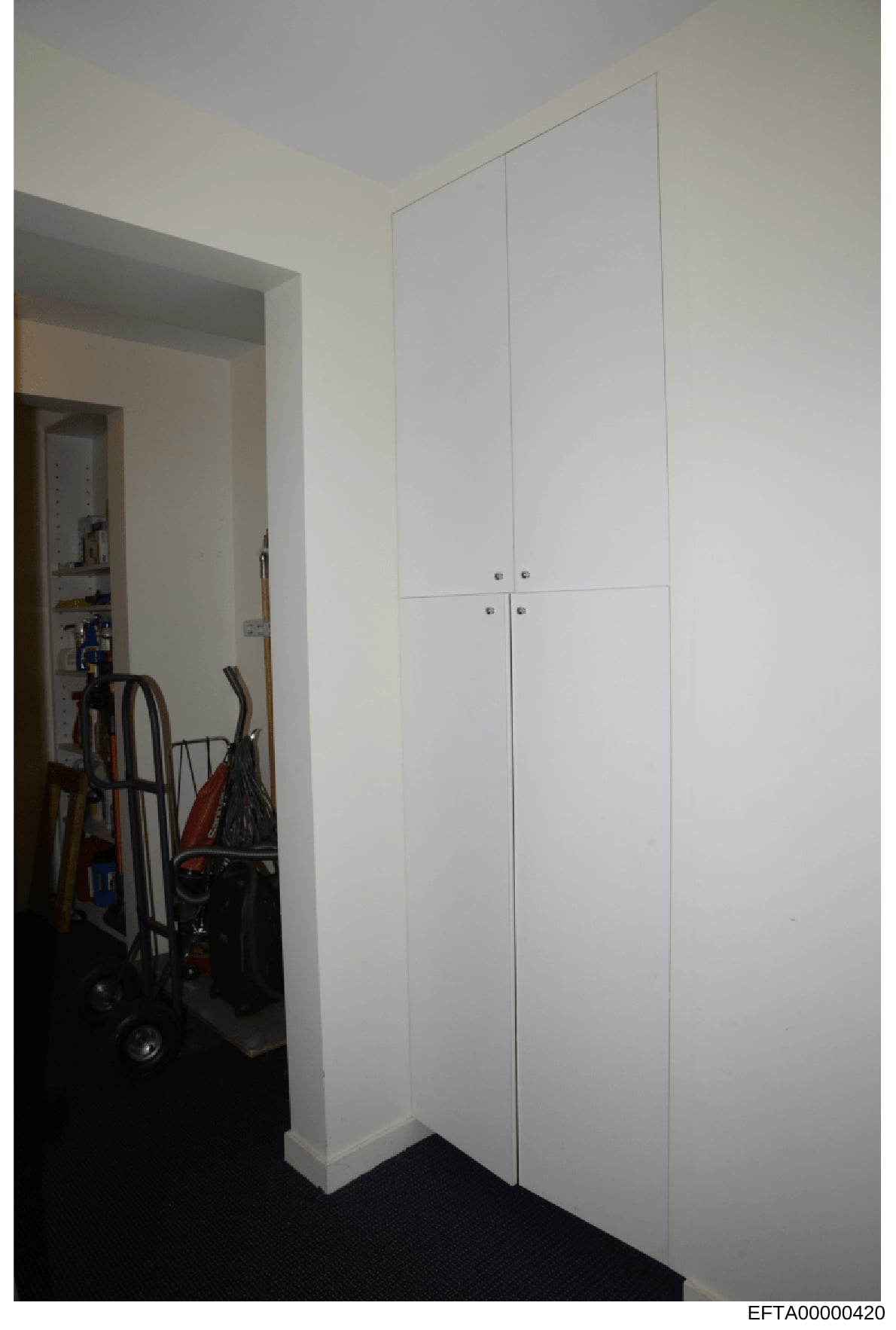 This is a photograph of an interior room showing a white four-door cabinet/closet unit and an open doorway leading to another room containing various household items and equipment. The image appears to be documentation of a residential property, poss