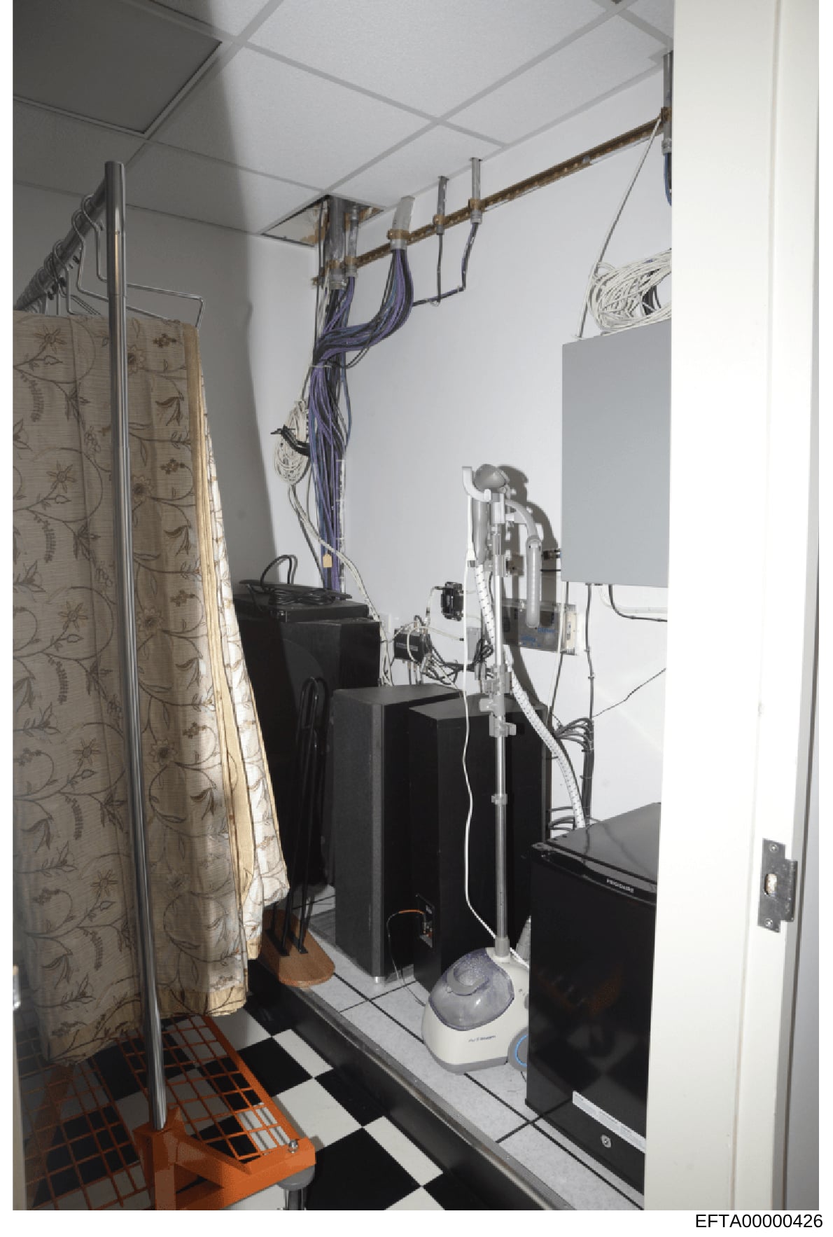 This is a photograph of an interior room space containing various electronic equipment, cables, and furnishings. The image shows a cluttered utility or technical space with black equipment boxes, overhead cable management, a patterned curtain, and ch