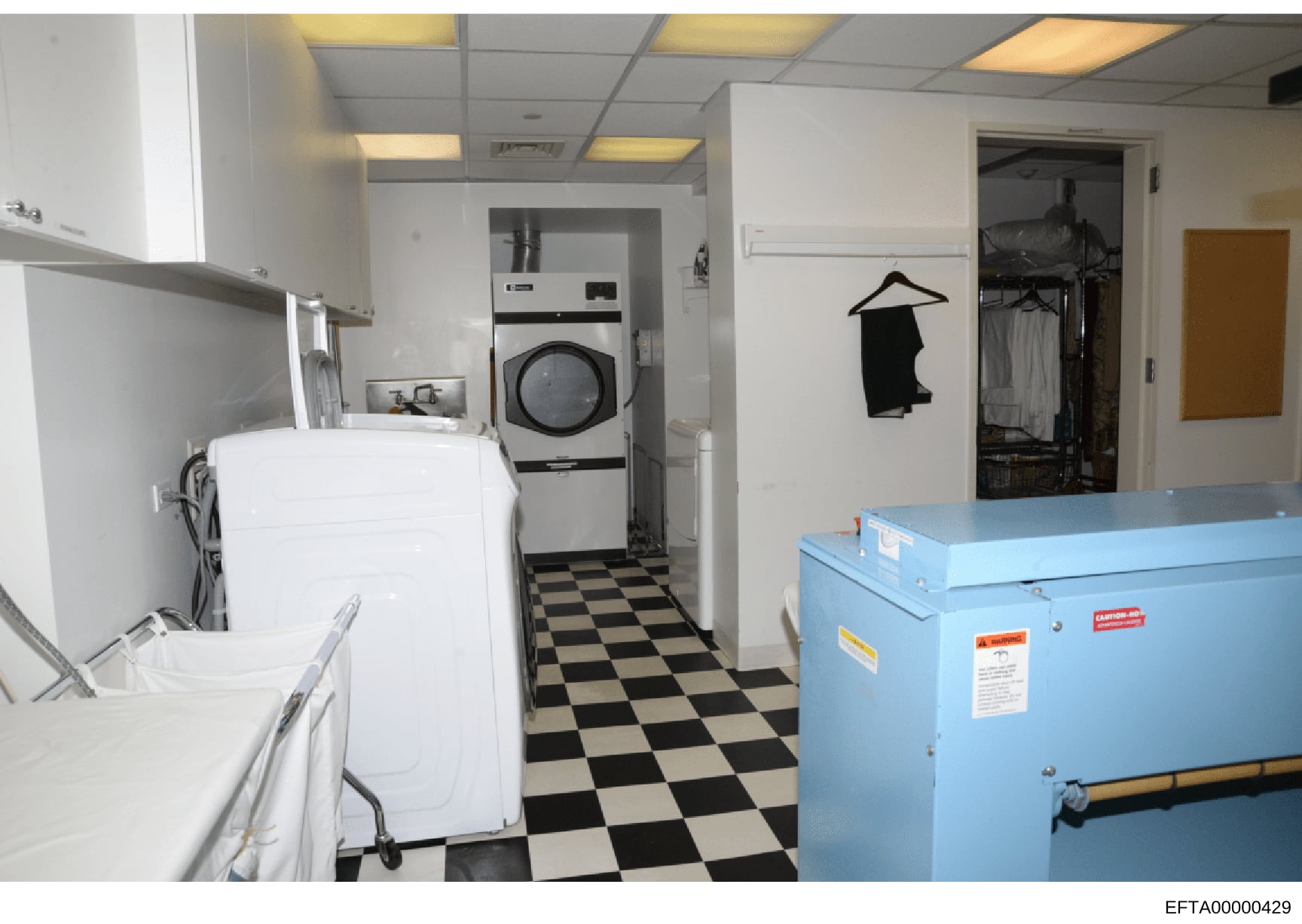 This photograph shows a commercial or institutional laundry room equipped with industrial-grade washing and drying equipment, including a white washing machine, a commercial dryer, and a large blue industrial washing or processing machine. The space