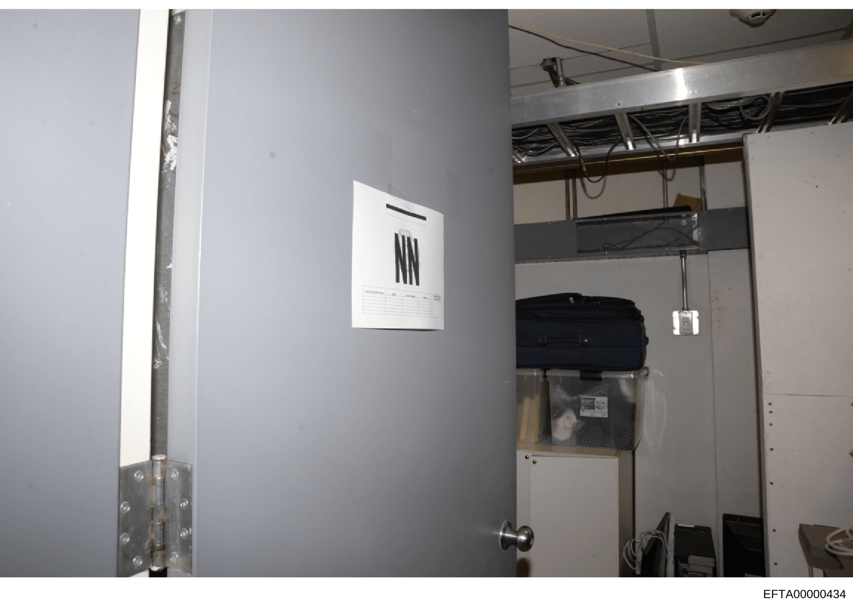 This is a photograph of an interior room, likely taken during the investigation, showing a closet or storage area with gray walls, exposed ceiling infrastructure, and stored items including a black bag/container. An evidence marker with tally marks i