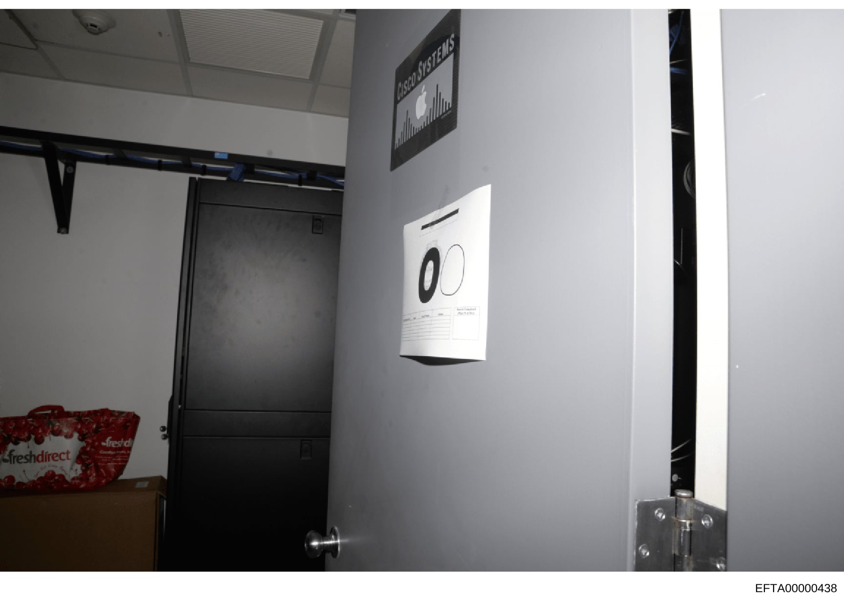 This is an evidence photograph showing the interior of a room or office space, featuring a black door, wall-mounted control system or alarm panel displaying circular indicators, shelving with a red 'FreshDirect' branded item, and various technical eq
