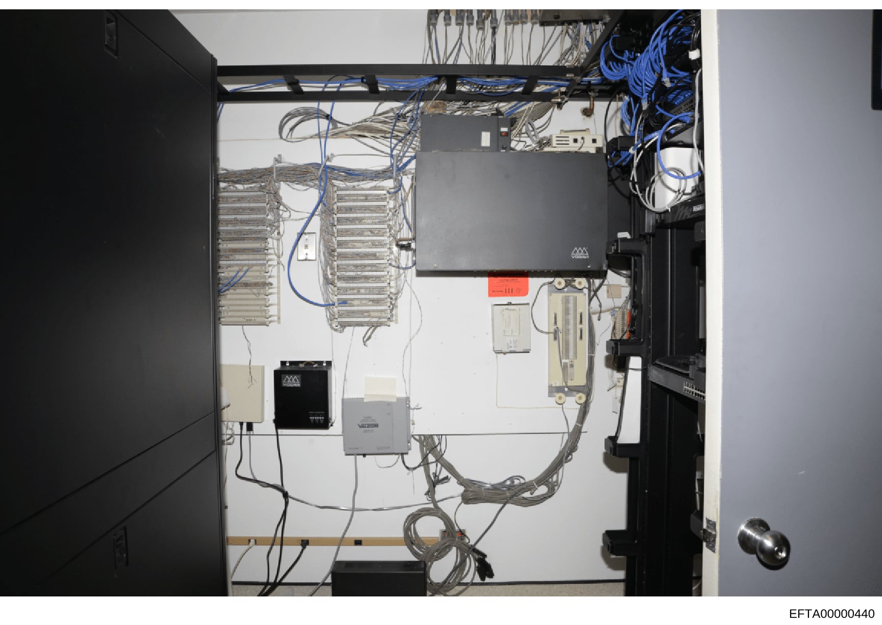 This is a photograph of an open server cabinet or network closet showing installed telecommunications and IT infrastructure. The image displays patch panels, network cabling (both blue and gray), power distribution equipment, black server/network dev