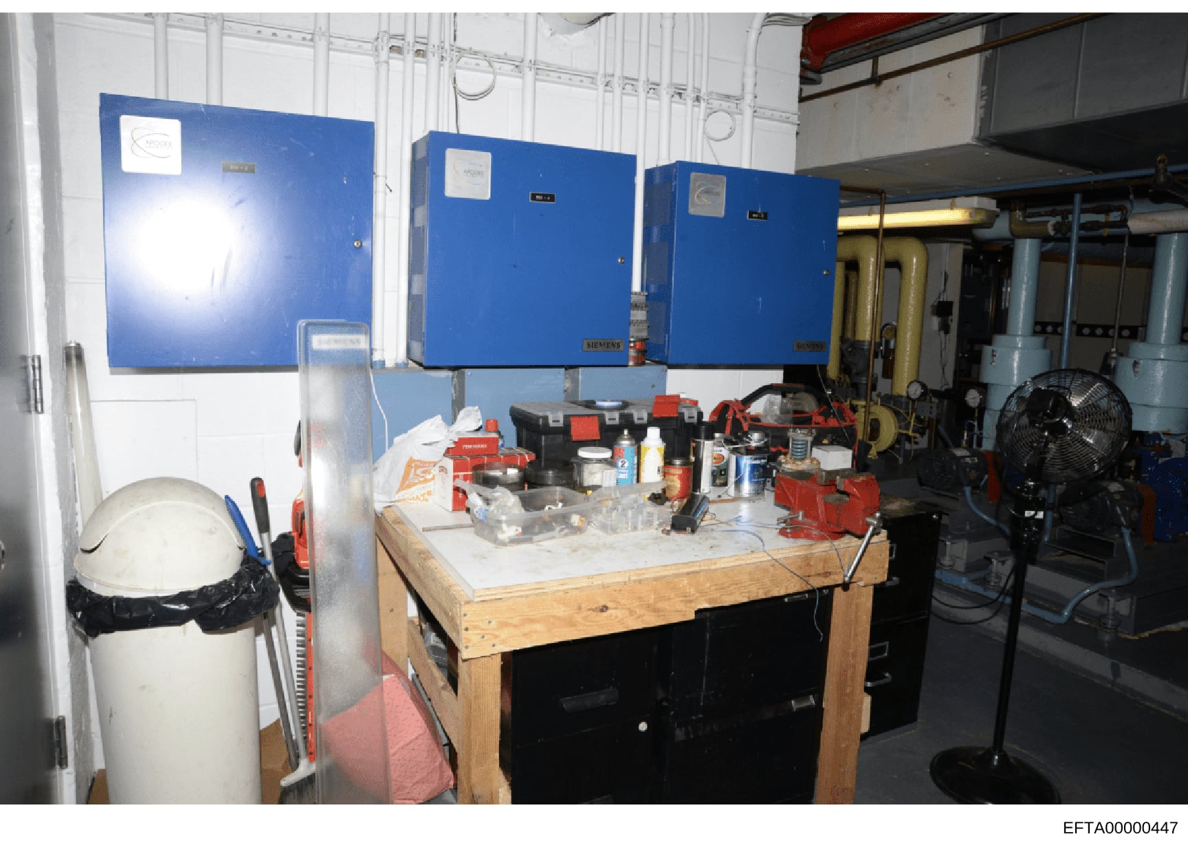 This photograph shows an interior workshop or utility space containing industrial electrical equipment (three large blue Siemens electrical boxes), a work bench with tools and supplies, a white cylindrical tank, safety equipment including a hard hat,