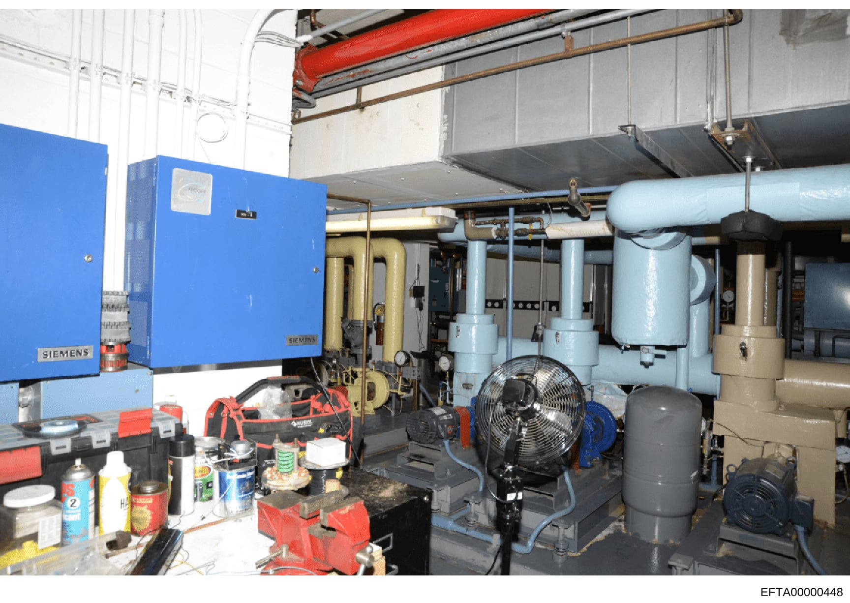 This photograph shows an industrial mechanical or utility room containing HVAC and building systems infrastructure. The image depicts a Siemens blue control panel, piping systems, fans, and various mechanical equipment in what appears to be a commerc