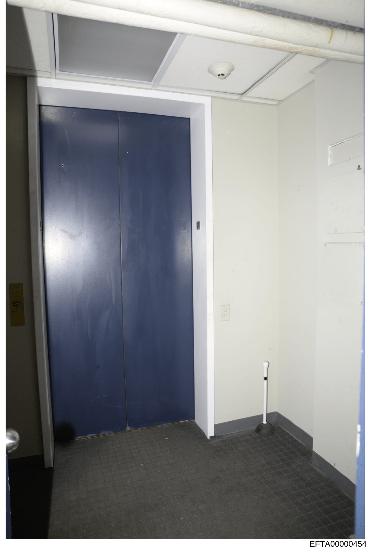 This is an evidence photograph depicting an interior hallway or corridor space, featuring a dark-framed elevator with navy blue doors and a white surround, cream-colored walls, drop ceiling with fire detection system, and dark flooring. The image app