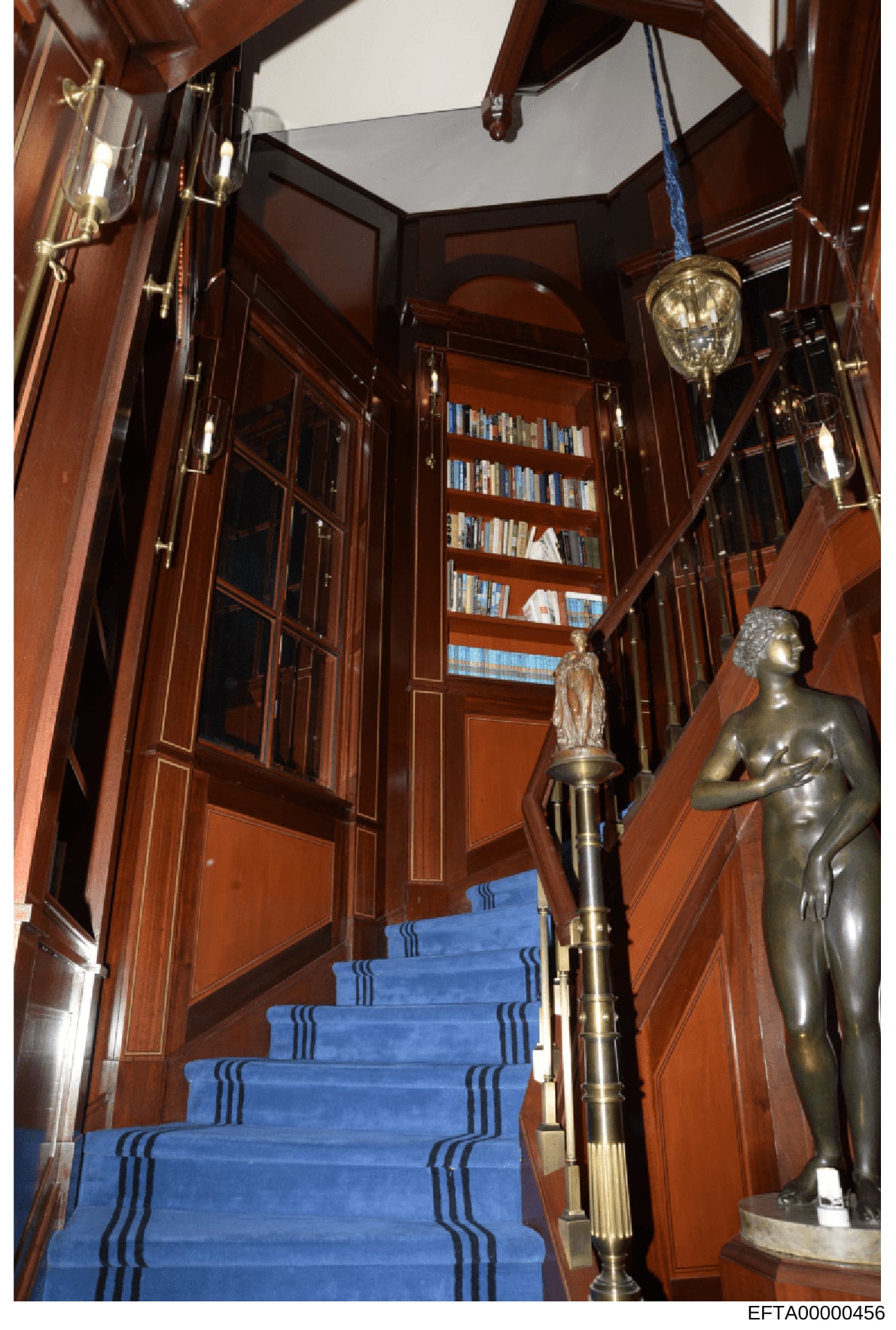 This is an evidence photograph showing the interior of what appears to be a high-end residential property, featuring an ornate wooden staircase with blue carpeting and decorative brass railings, flanked by classical nude bronze sculptures and an exte