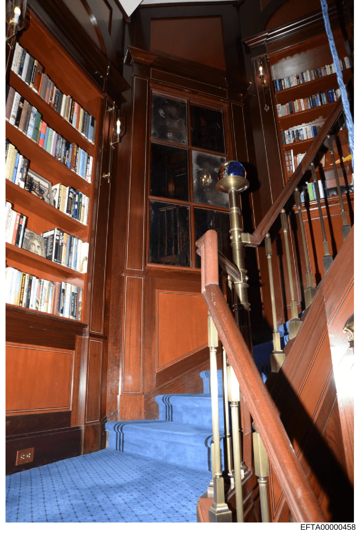 This is a photograph of an interior room featuring floor-to-ceiling mahogany bookshelves stocked with books and media on both sides, a large window with interior shutters in the center, blue carpeting, and a brass handrail. The image appears to be fr