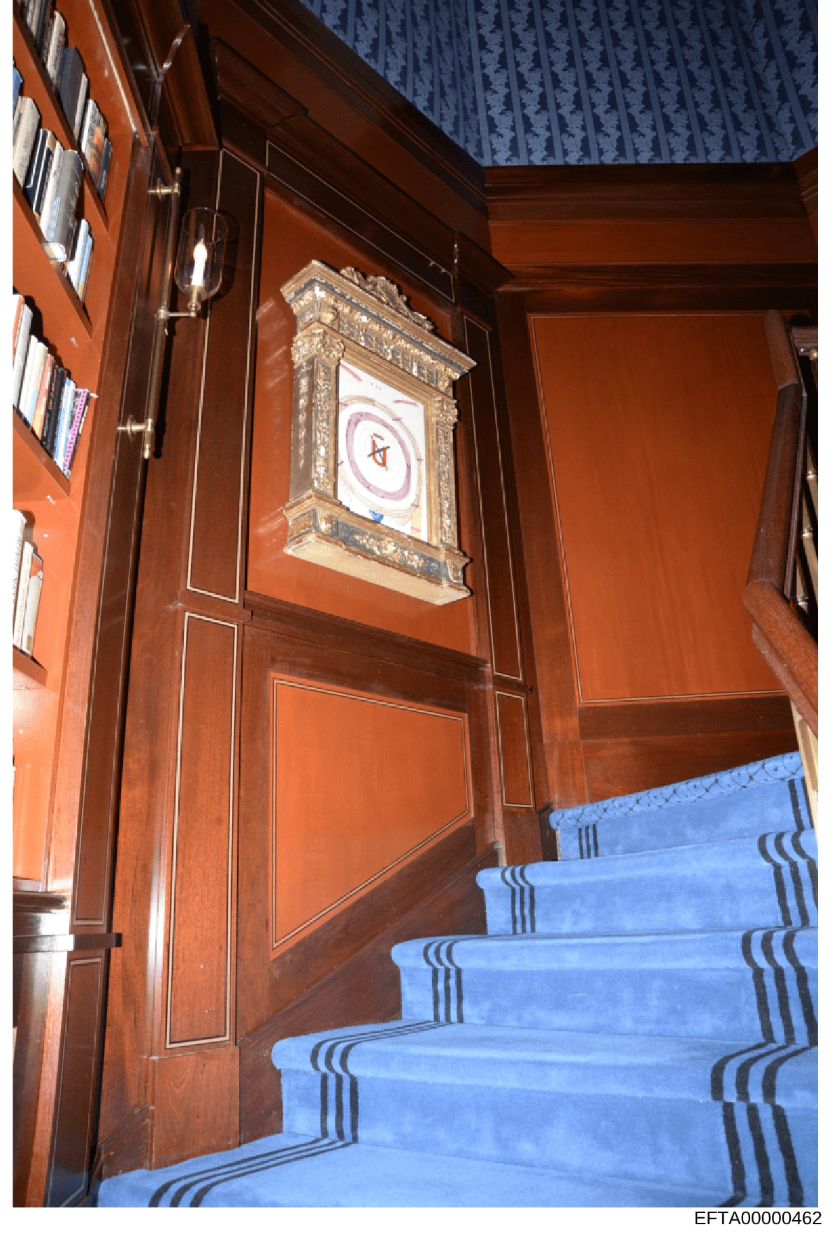 This is an evidence photograph showing the interior of a residential room featuring rich wood paneling, a blue carpeted staircase with decorative striping, and a framed artwork with an ornate gilded frame mounted on the wall. The photograph documents