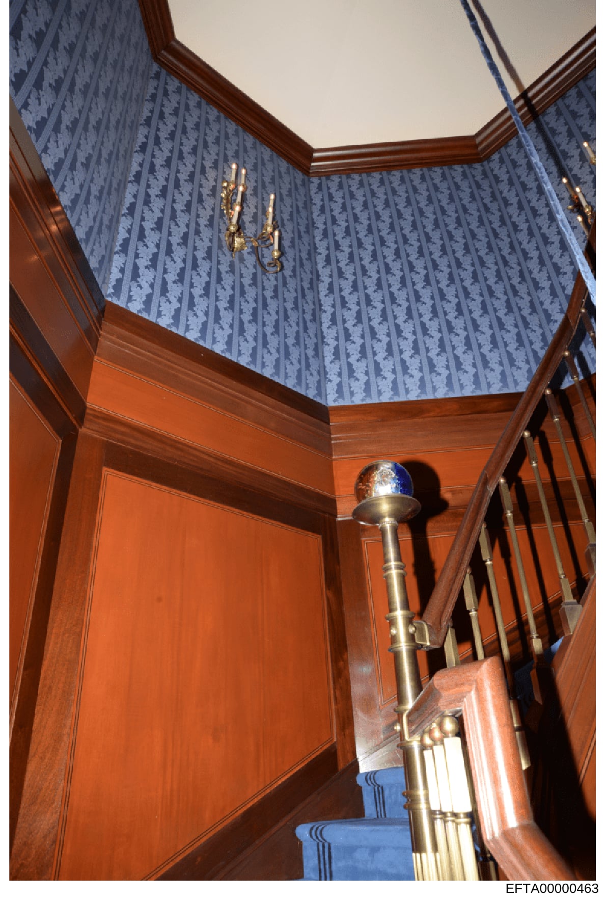 This is an evidence photograph showing the interior of a residential stairwell, likely from one of Jeffrey Epstein's properties. The image displays the architectural details including rich wood paneling, blue damask wallpaper, ornate brass sconces, a