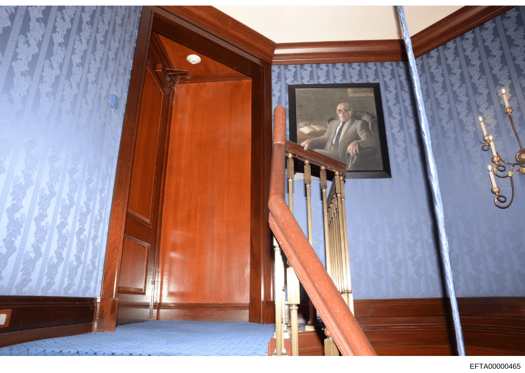 This is an evidence photograph documenting an interior hallway and upper landing area featuring elegant wood-paneled doors, blue damask wallpaper, brass wall sconces, and a framed portrait of a man in professional attire. The photograph appears to be