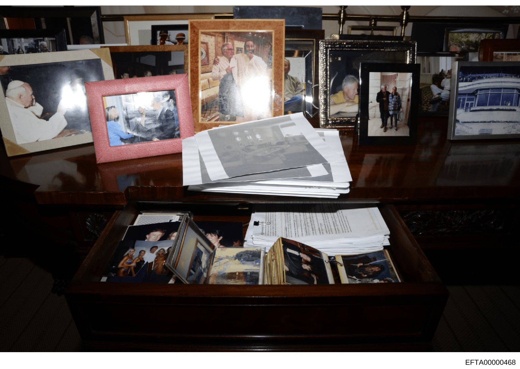 This is a photograph of a wooden desk displaying multiple framed photographs, loose photographs, and stacked documents. The image appears to be evidence documentation from the investigation, showing a collection of personal items and papers arranged 