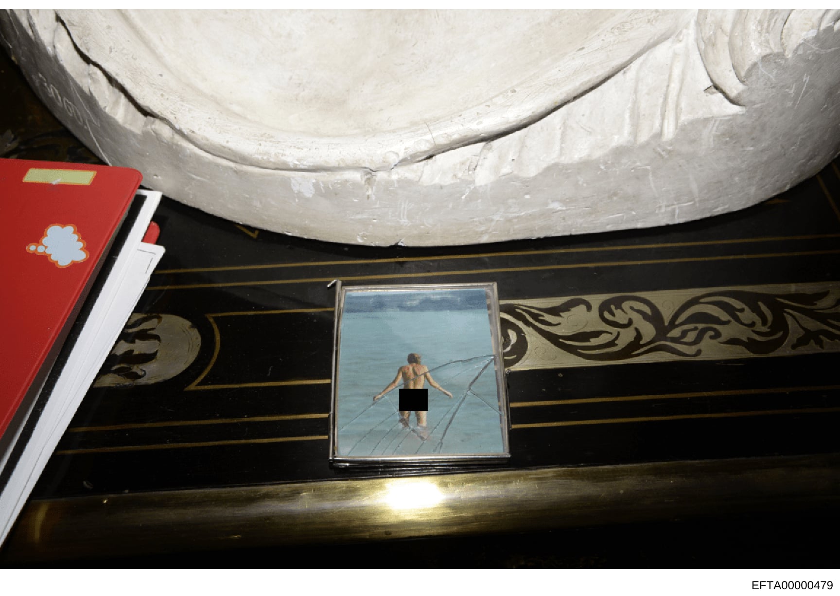 This is a documentary photograph showing a framed picture discovered during an investigation search, likely of an Epstein property. The photo depicts a male figure in water and is mounted in an ornate black frame with gold decorative trim. The image 