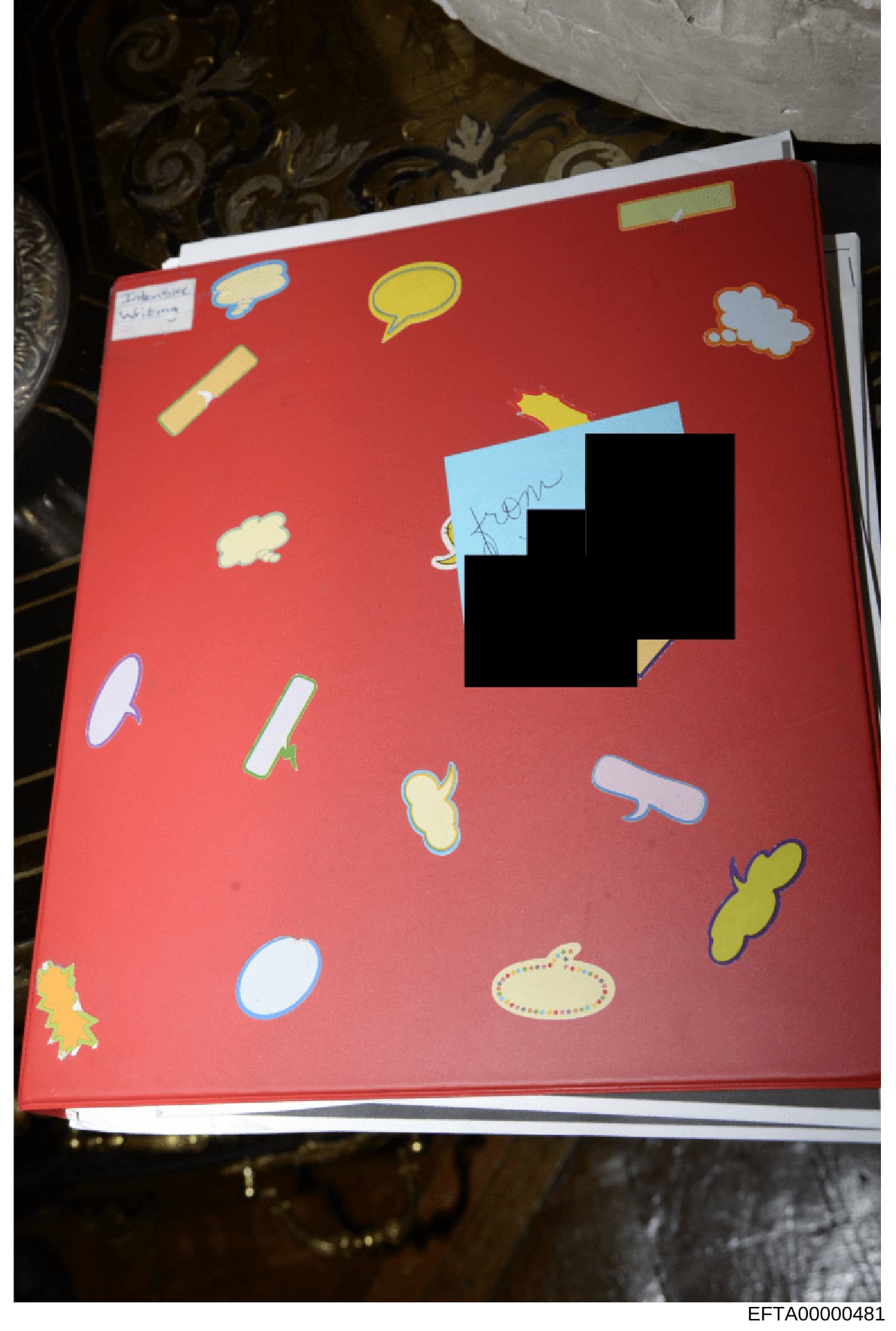 This is a photograph of a red-covered notebook or journal decorated with various colorful stickers and handwritten markings. A significant portion of the cover is obscured by black redaction blocks. The item is photographed against a patterned backgr
