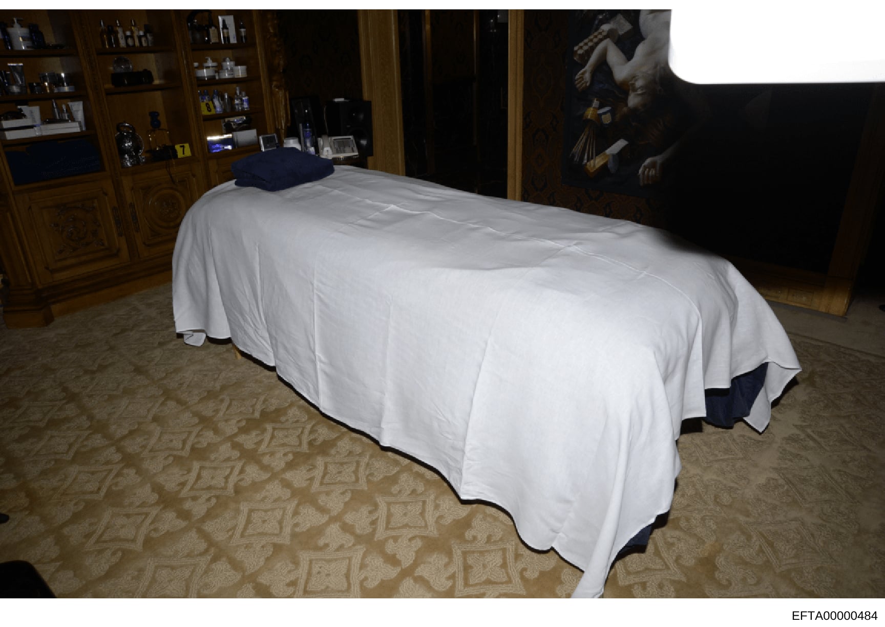 This is an evidence photograph showing a bedroom interior with a bed covered in white linens and a dark blue pillow, wooden cabinetry with decorative items, an ornate wooden door frame, patterned carpeting, and artwork on the wall. The photograph app
