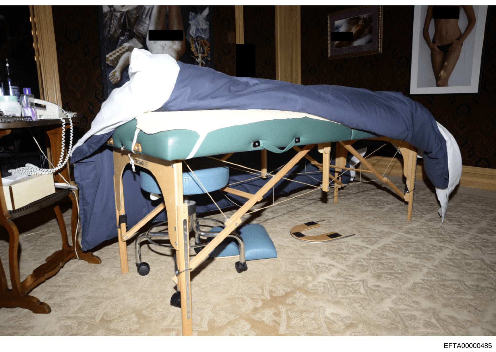 This photograph shows the interior of a bedroom containing a portable massage table with teal and blue cushioning, wooden frame, and blue draping. The room includes a wooden side table with personal items, artwork on the walls, and various furnishing