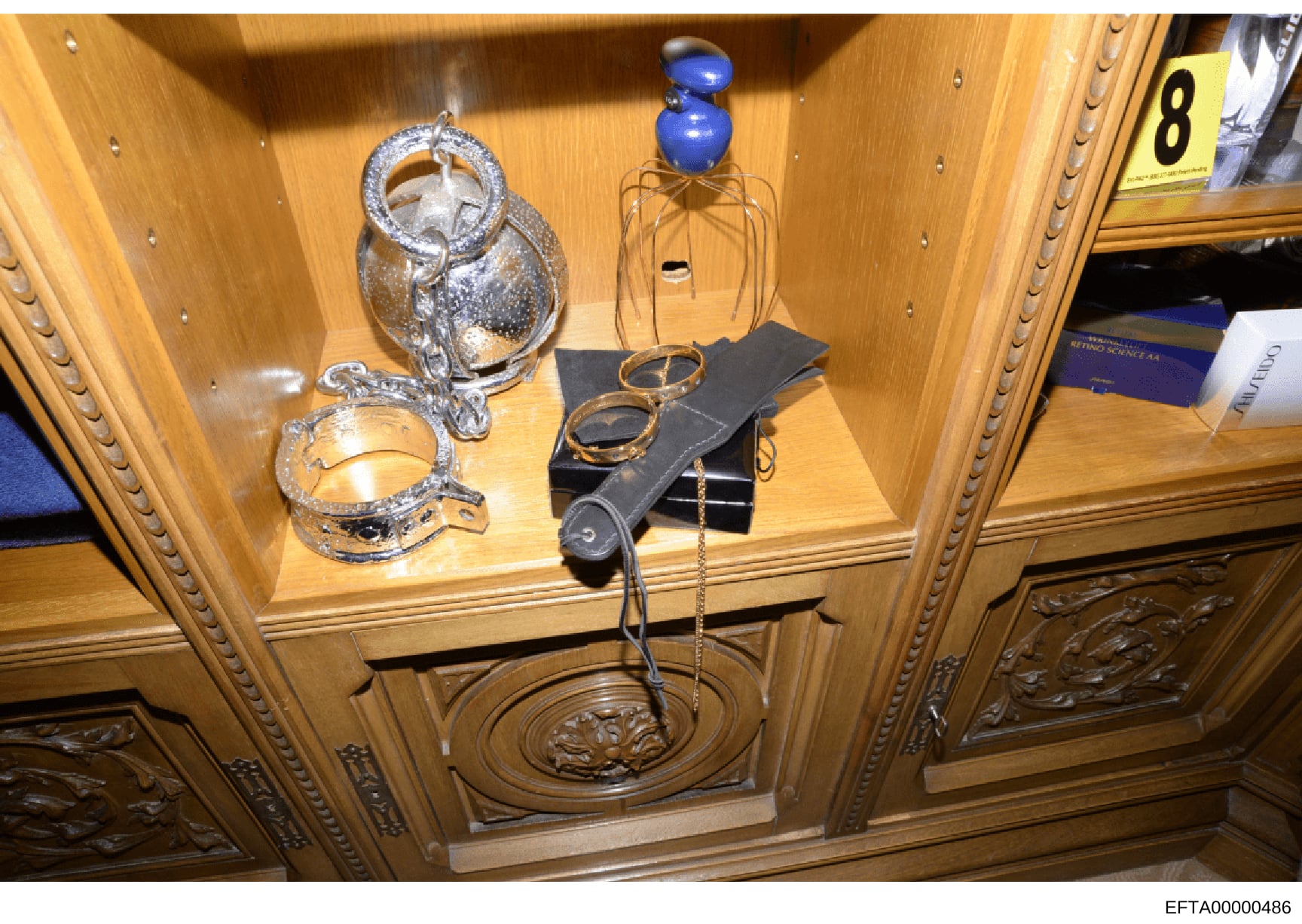 This is an evidence photograph showing the interior of a wooden cabinet or display case containing various decorative items and jewelry. The items include ornate silver-colored containers, blue decorative objects, gold-toned wire sculptures, and blac
