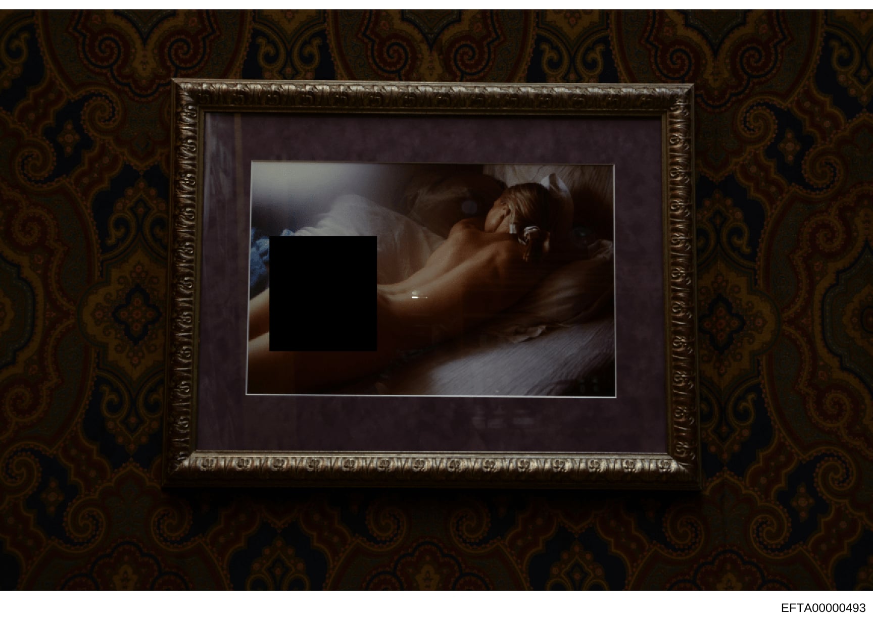 This is an evidence photograph showing a framed picture hung on a wall with distinctive ornate patterned wallpaper in gold and dark tones. The photograph within the frame contains a large black redaction box obscuring the primary content. The image a