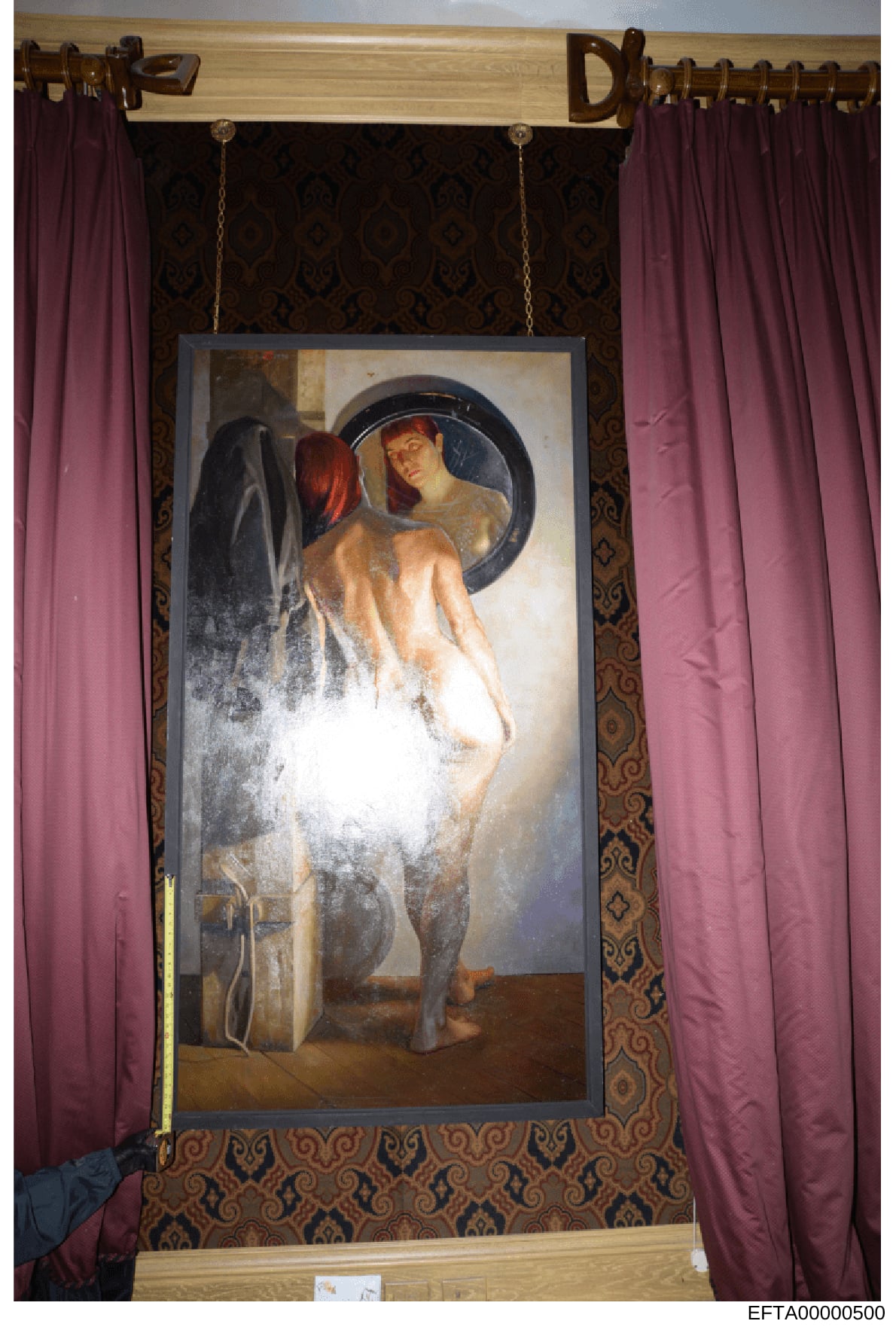 This is a photograph of a framed painting displayed on a wall, showing classical or allegorical artwork featuring a female figure with red hair viewing her reflection in a circular mirror. The painting is mounted in an ornate wood frame with decorati