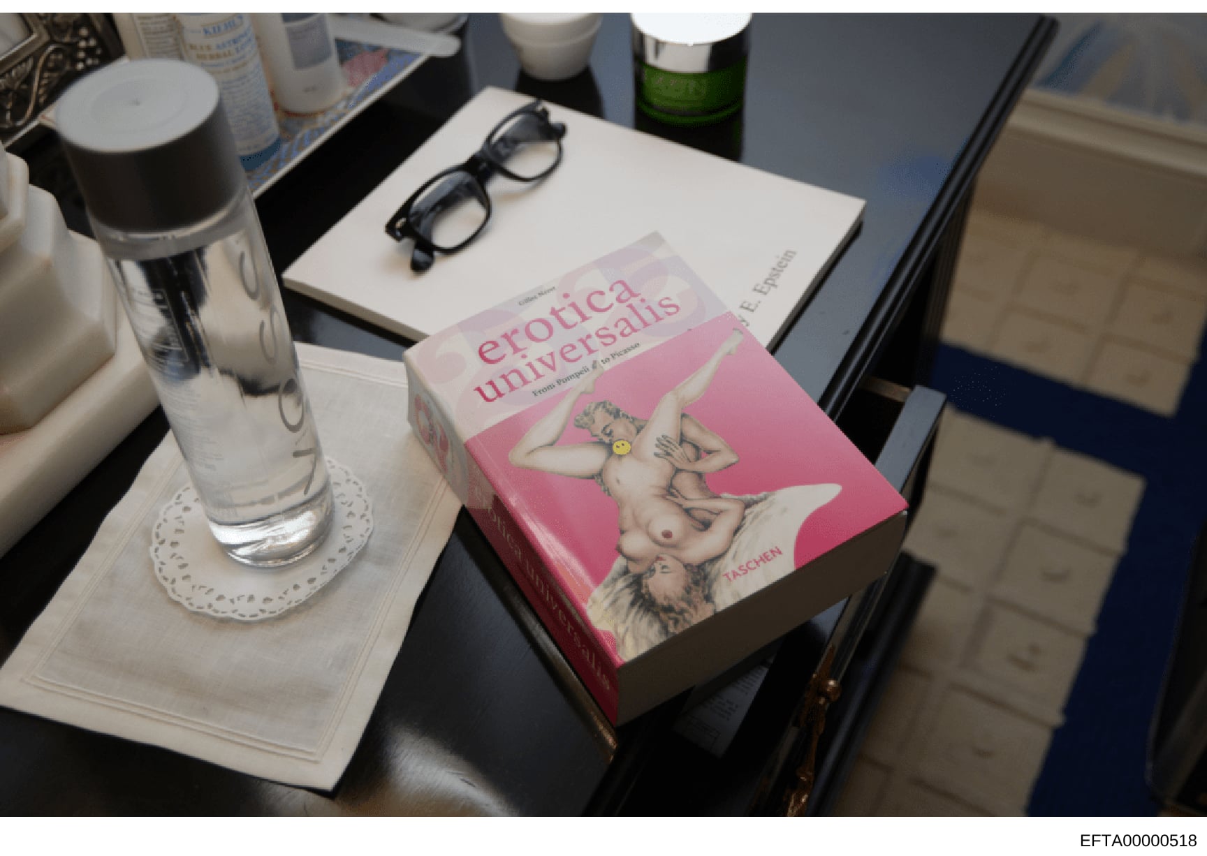 This is an evidence photograph showing a pink-covered book titled 'Erotica Universalis' (published by Taschen) displayed on a nightstand or table alongside personal items including eyeglasses, a water bottle, toiletries, and a green bottle. The photo