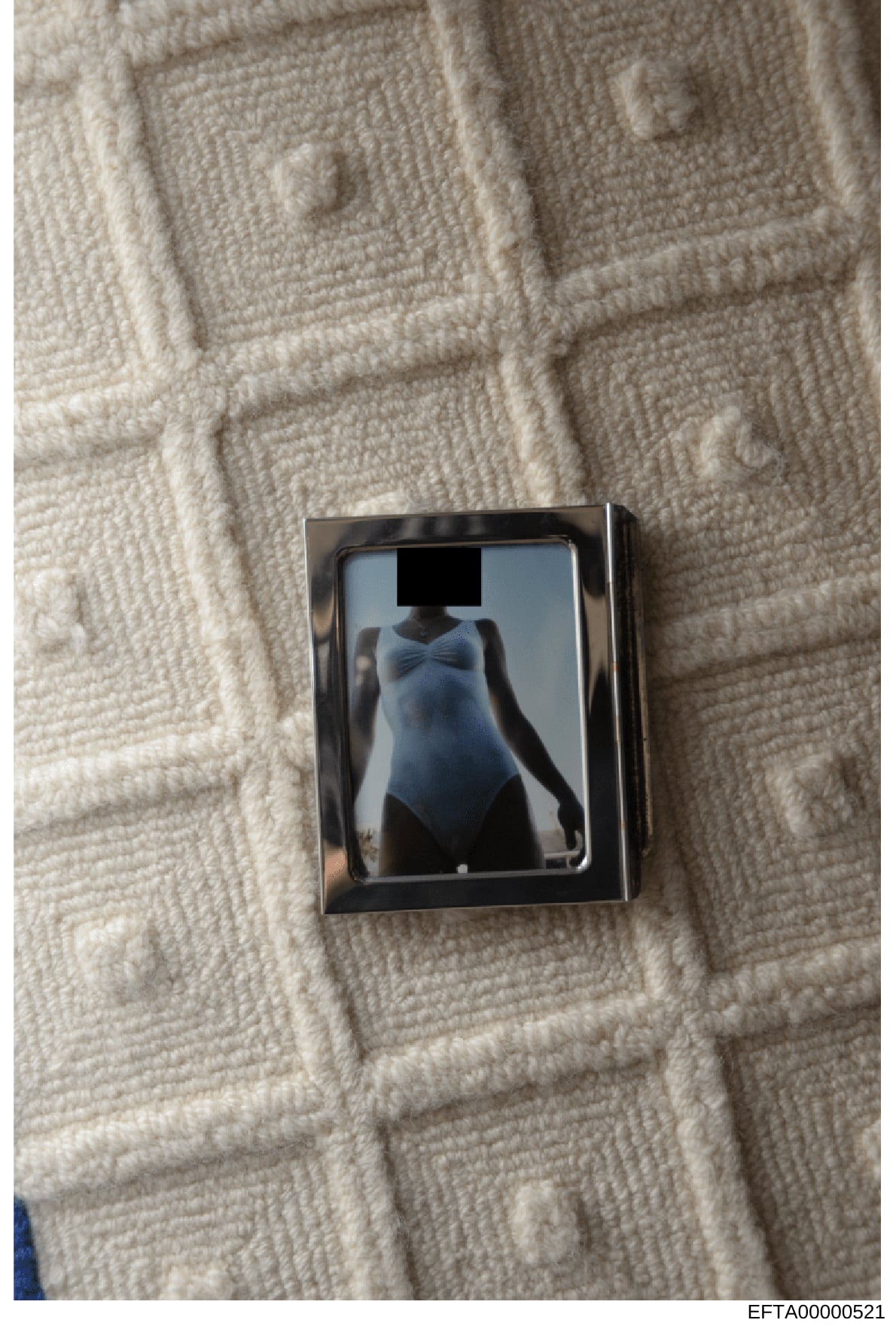 This is a photograph of a framed image placed on a cream-colored quilted or textured fabric surface. The image within the frame shows a figure in a light blue/gray garment with the face redacted by a black rectangle. The photo appears to be evidence